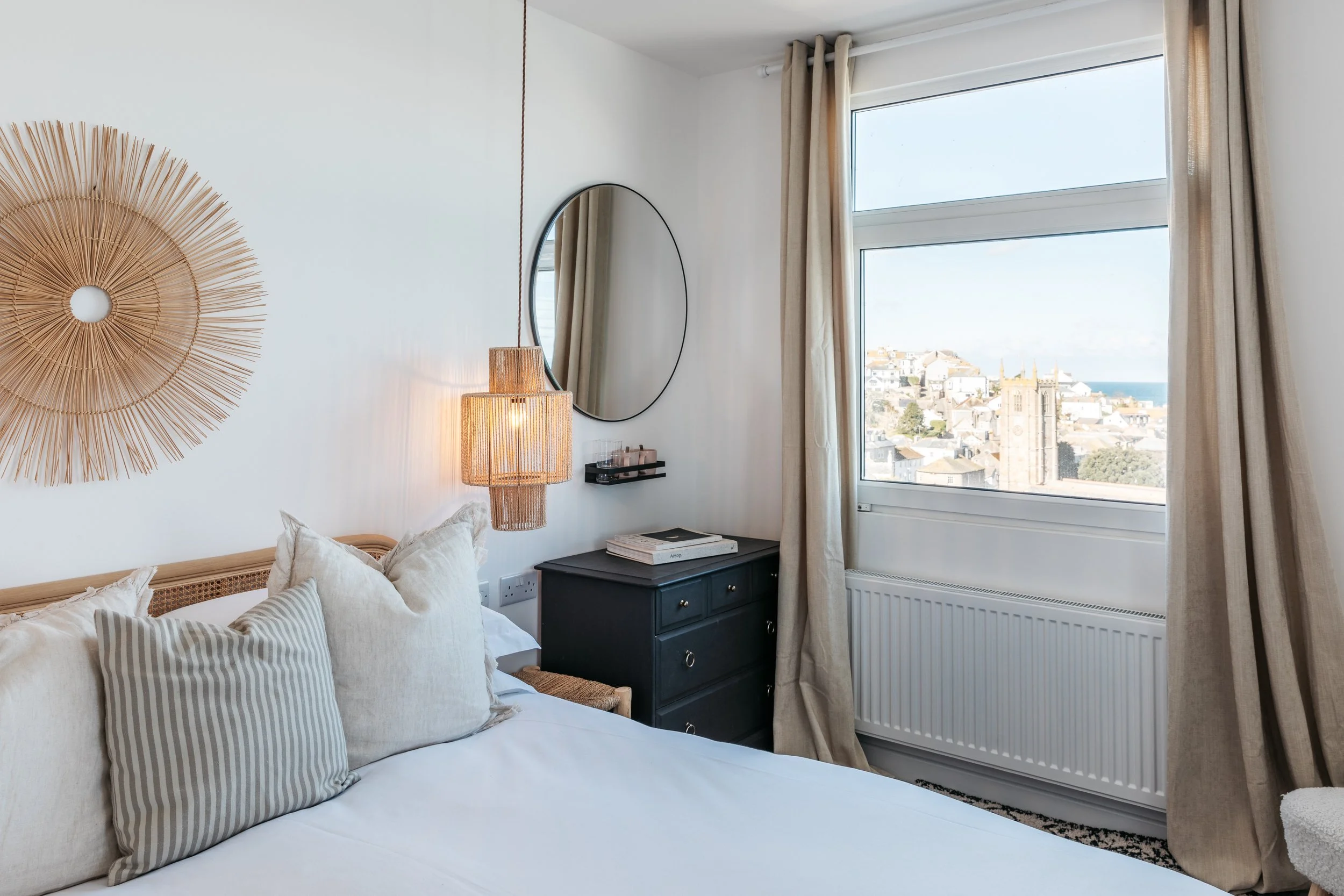 A cozy bedroom with a bed featuring striped and solid pillows, a black dresser with stacked books on top, a round mirror on the wall, a woven hanging decoration, a hanging lamp, and a large window with curtains showing a cityscape and a church tower