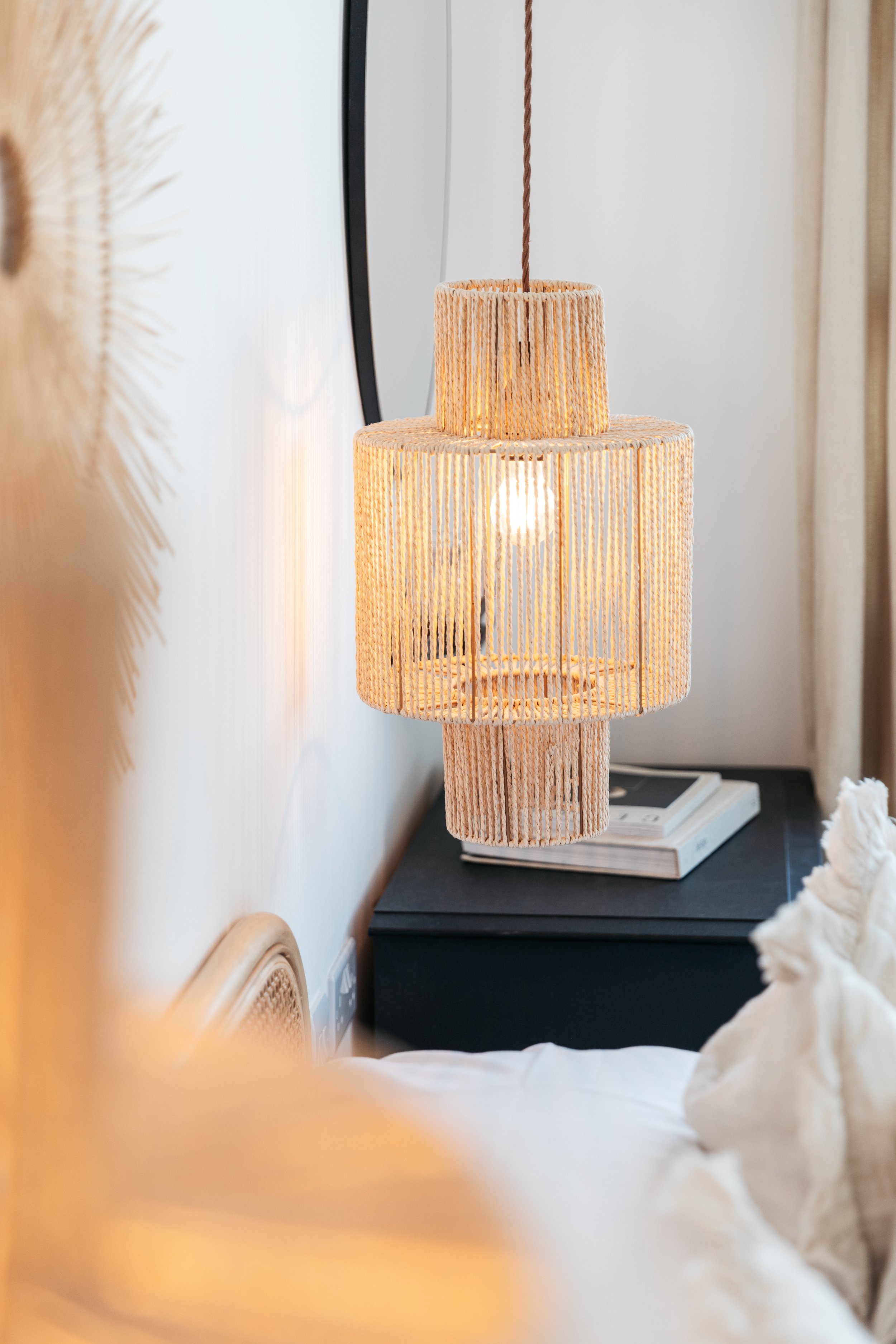 A hanging woven lampshade with a lightbulb inside, glowing, in a bedroom next to a bed and a black nightstand with books.