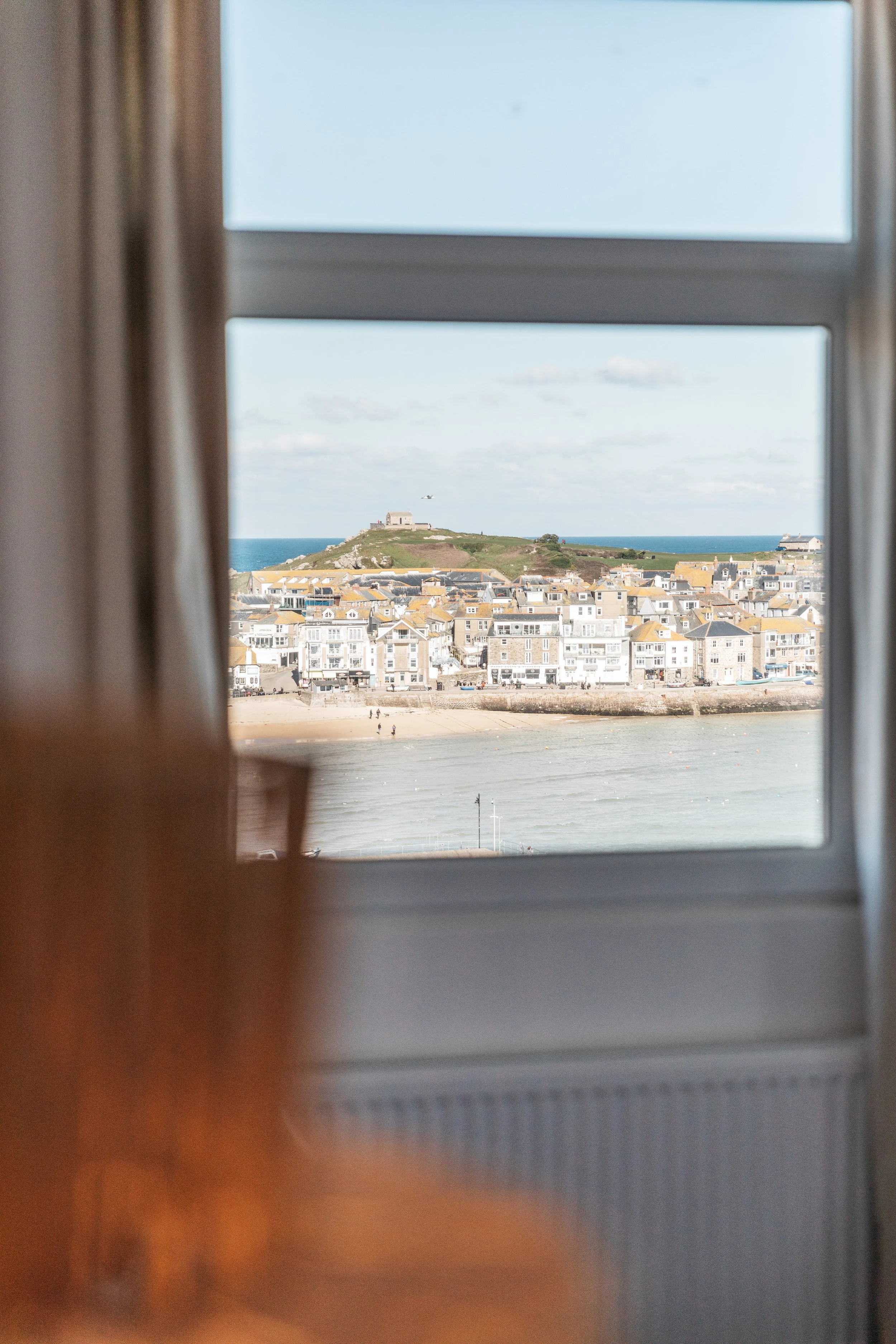 View of a coastal town with buildings, beach, and a hill with a structure on top, seen through a window.
