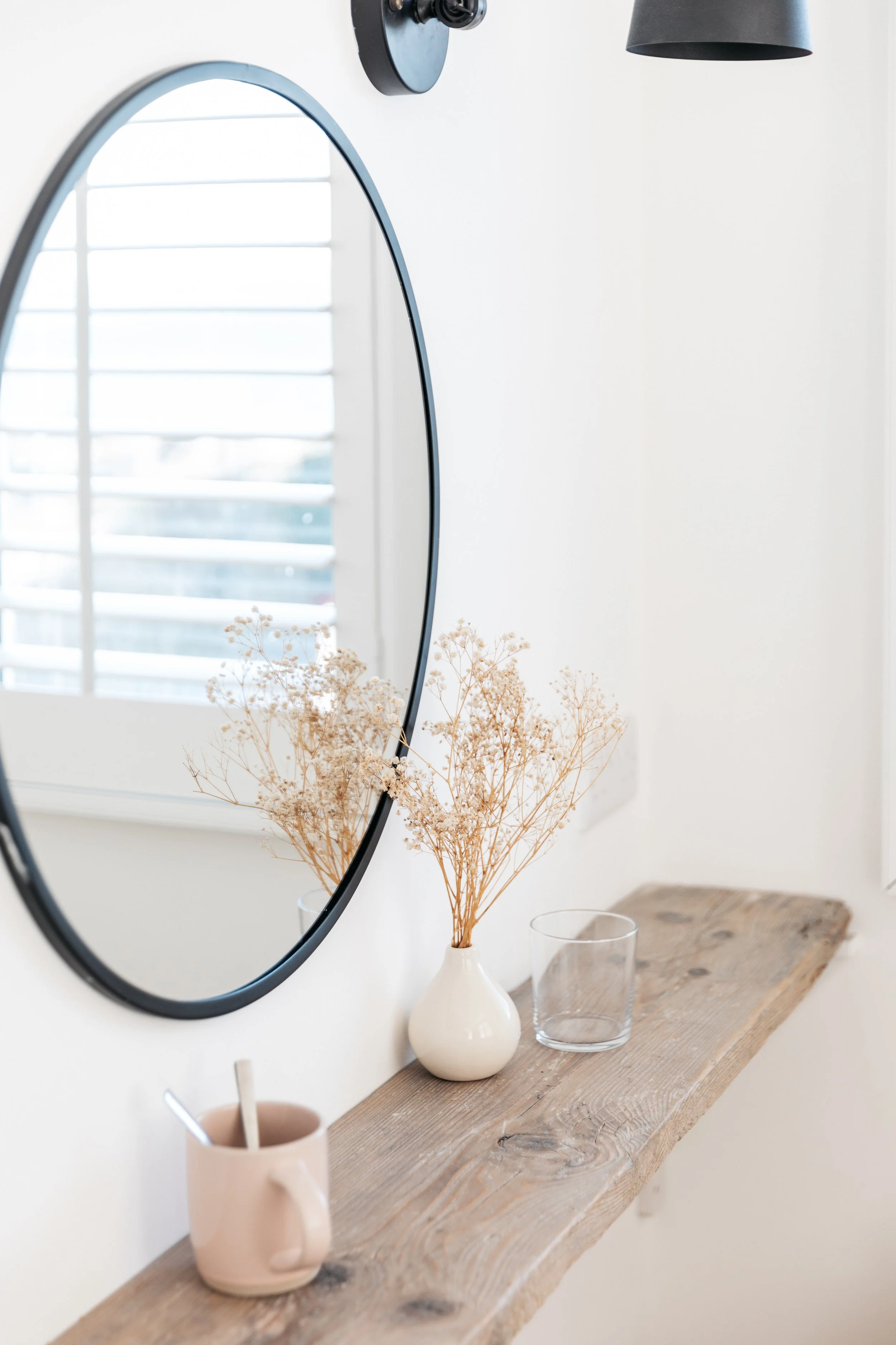 Decorative bathroom shelf with a round mirror, a vase with dried flowers, a glass, and a mug with utensils, in front of a window with blinds.