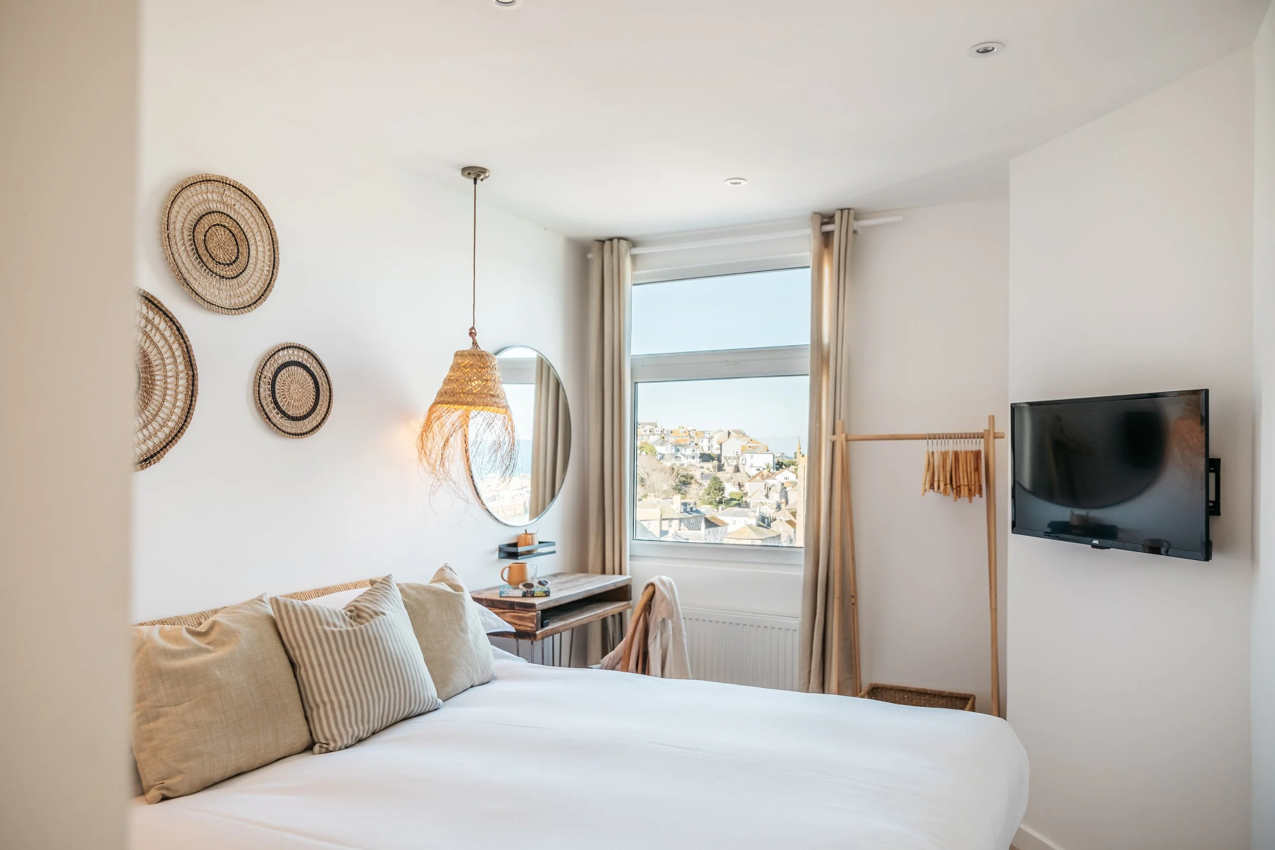 Cozy bedroom with a large bed, beige pillows, a round mirror, wicker wall decor, a window with city view, a blanket rack, and a flat-screen TV on the wall.
