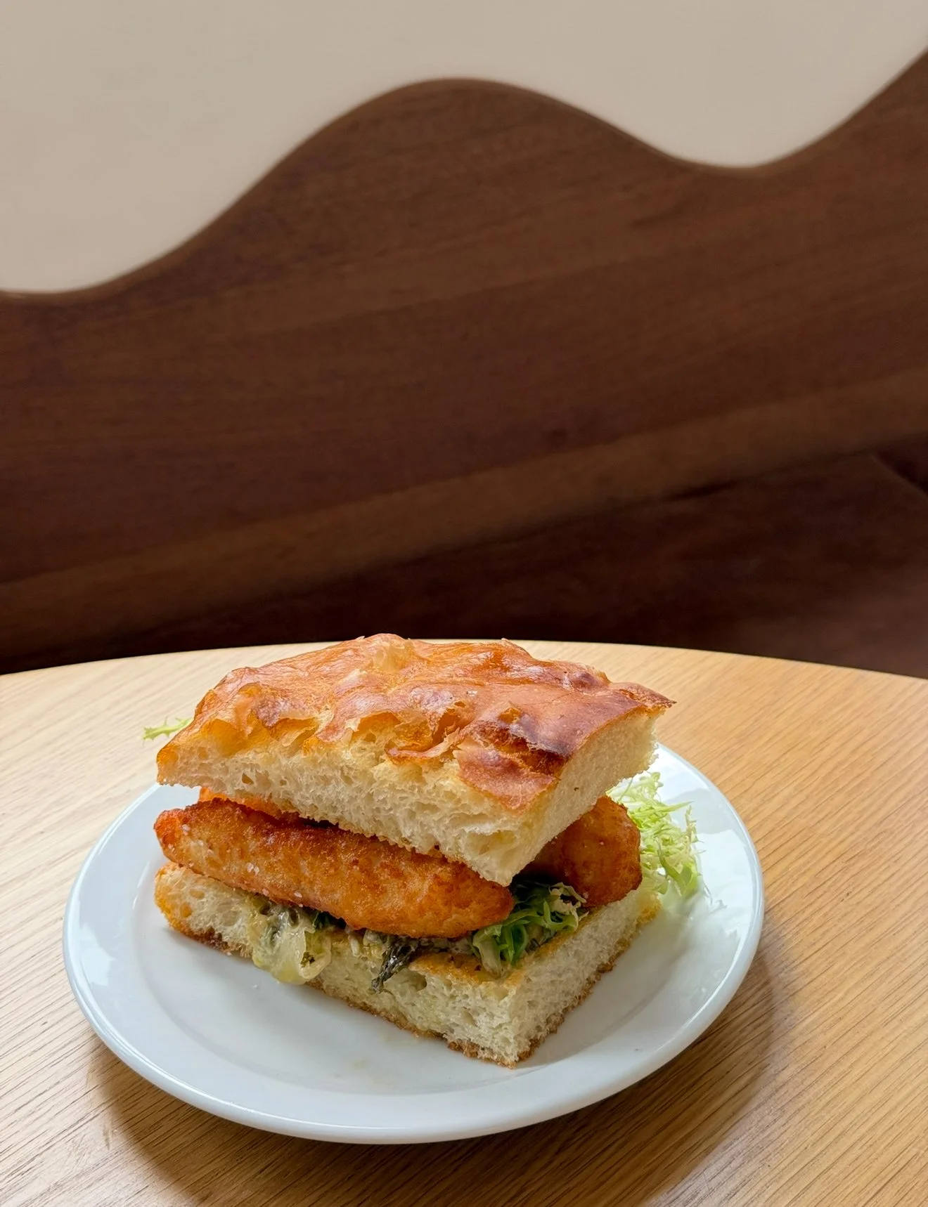 A sandwich on a white plate with a piece of crispy fried chicken, lettuce, and a slice of bread, with a wooden headboard in the background.