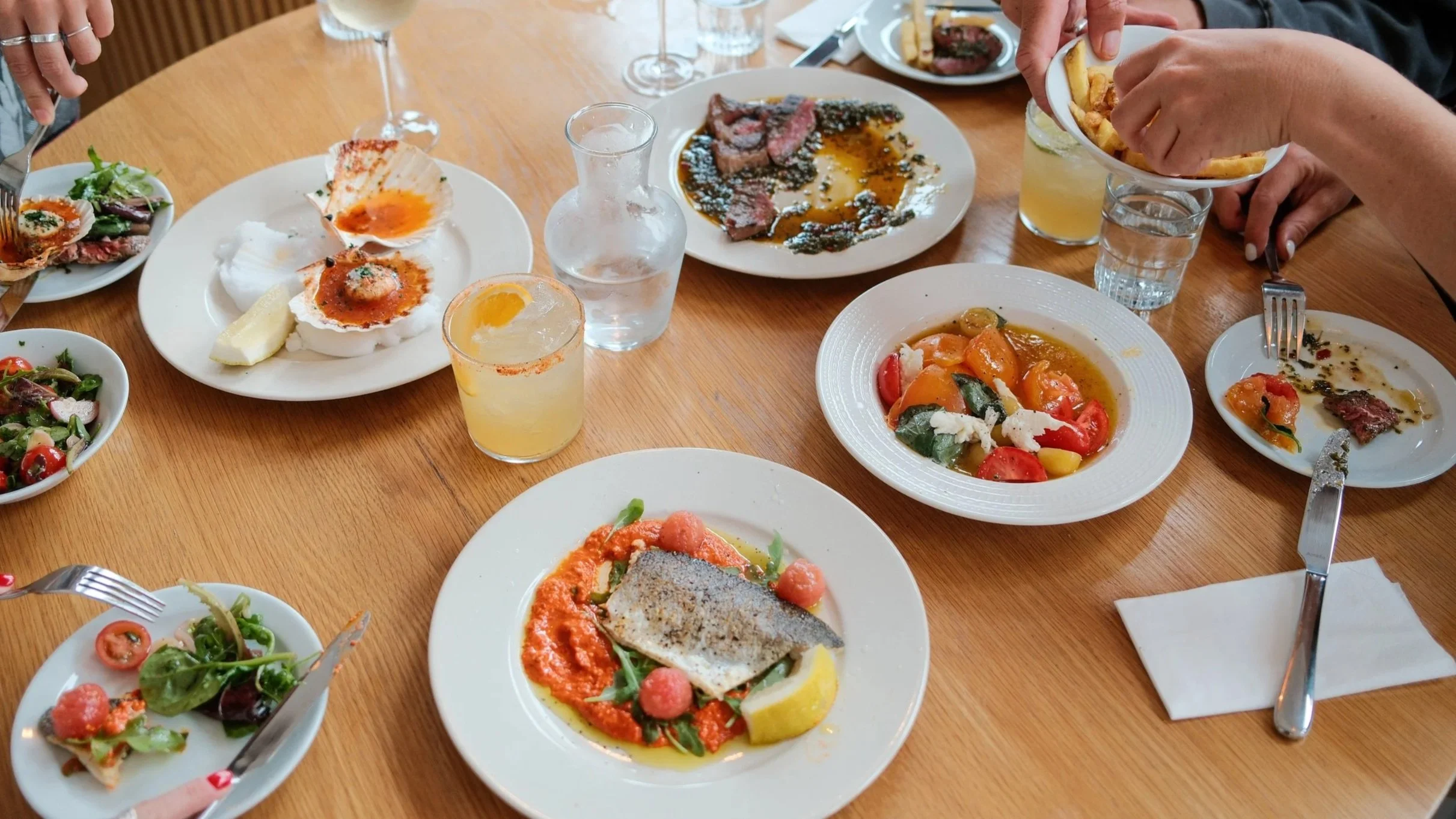 A variety of dishes on a wooden table including salads, grilled meats, and vegetables, with some beverages and utensils visible.