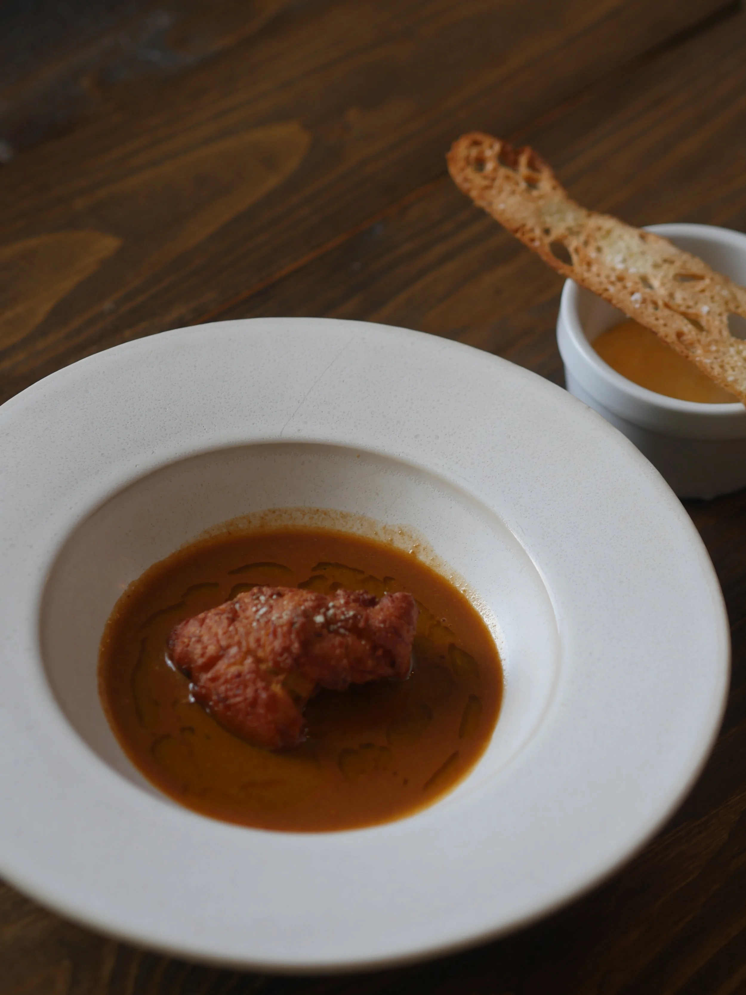 A bowl of soup with a piece of fried chicken floating in it, accompanied by a small dish of sauce with a crispy breadstick.