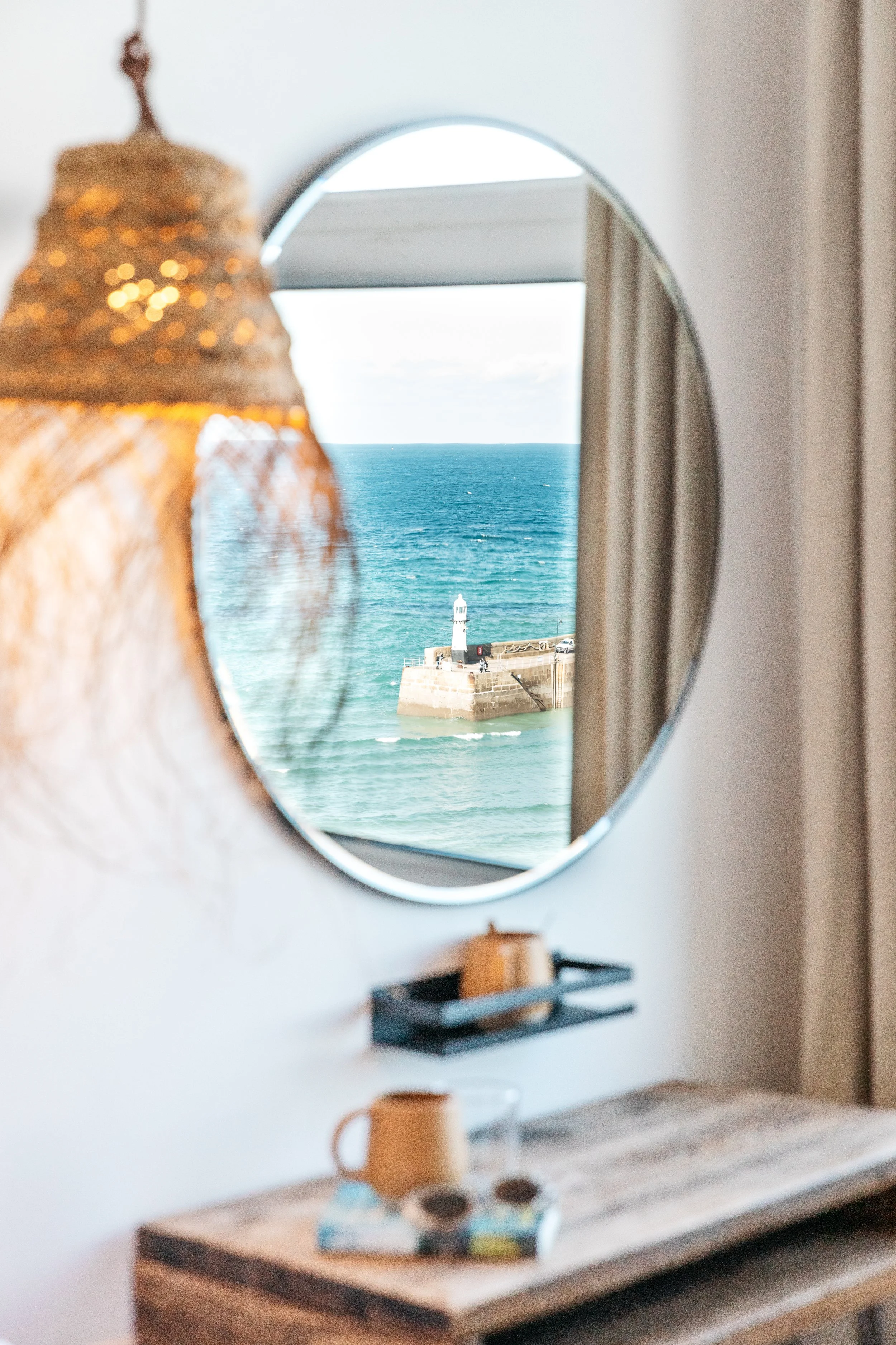 A round mirror on a white wall reflecting a view of the ocean with a lighthouse on a pier.