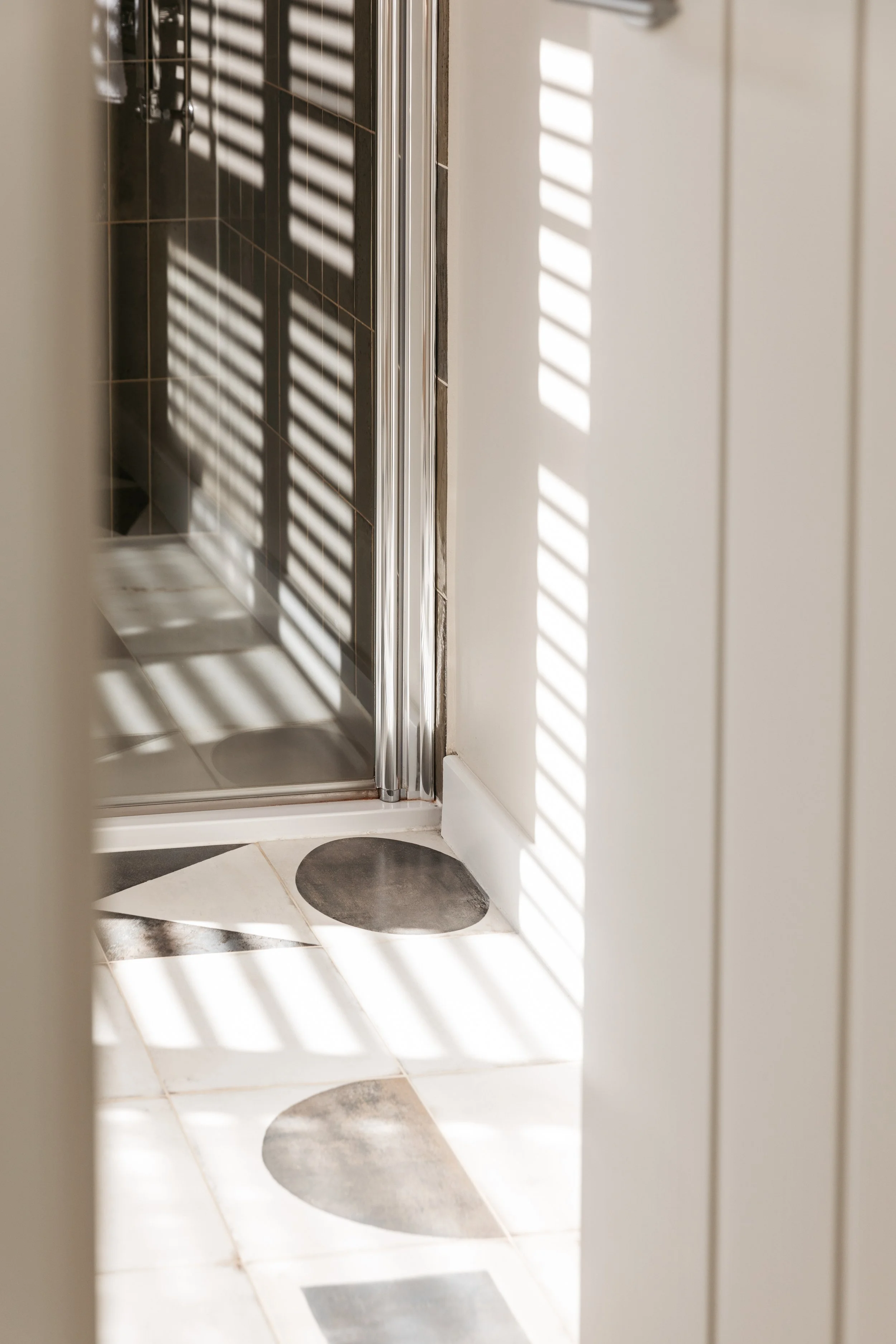 A view of a bathroom floor with patterned tiles and a shower with glass door, seen through a partially open door with sunlight and shadow stripes.