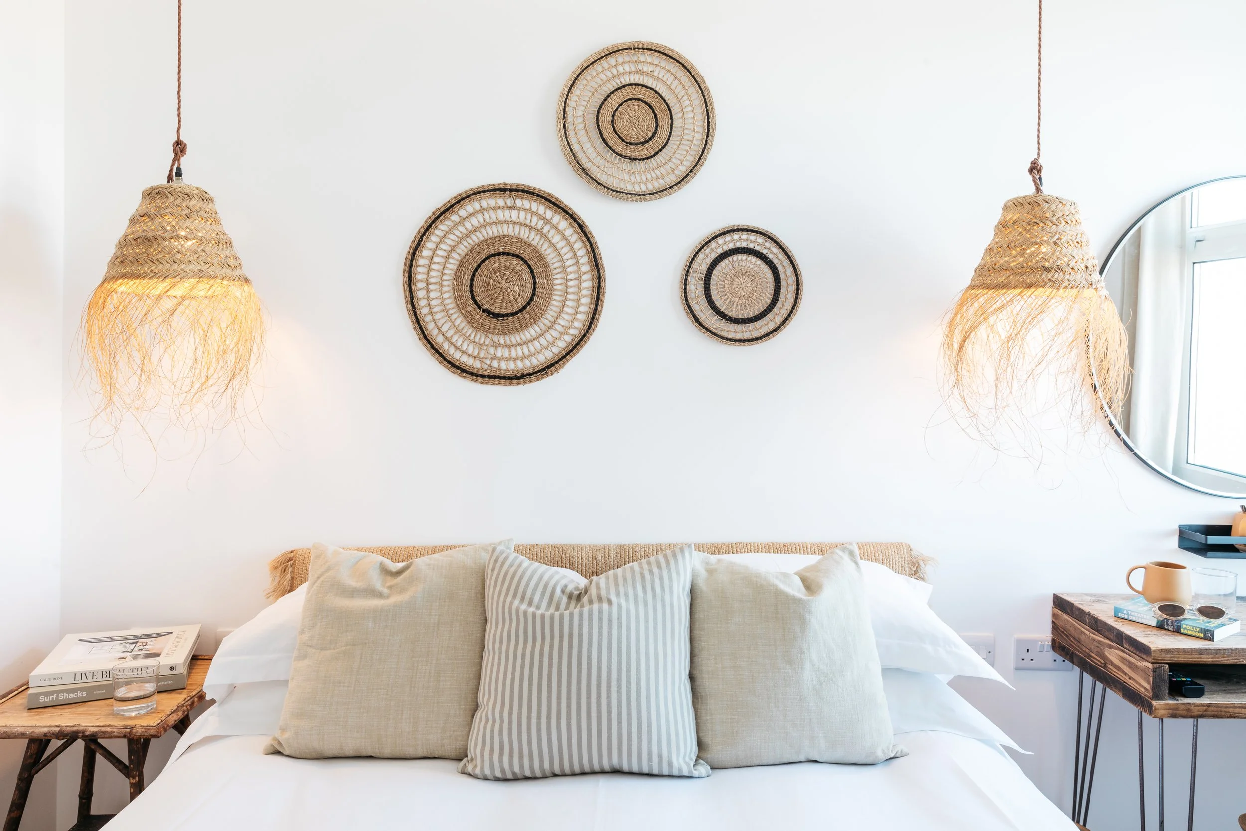 A bedroom with a bed, beige pillows, woven wall decor, hanging woven lamps, a round mirror, and a wooden bedside table with books, a glass of water, a mug, and decorative items.