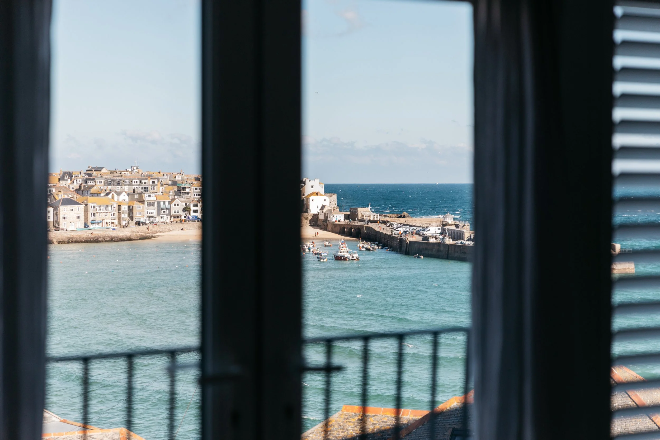 View of a harbor with boats, a sandy beach, and a town with pastel-colored buildings, seen through an open window with black shutters.