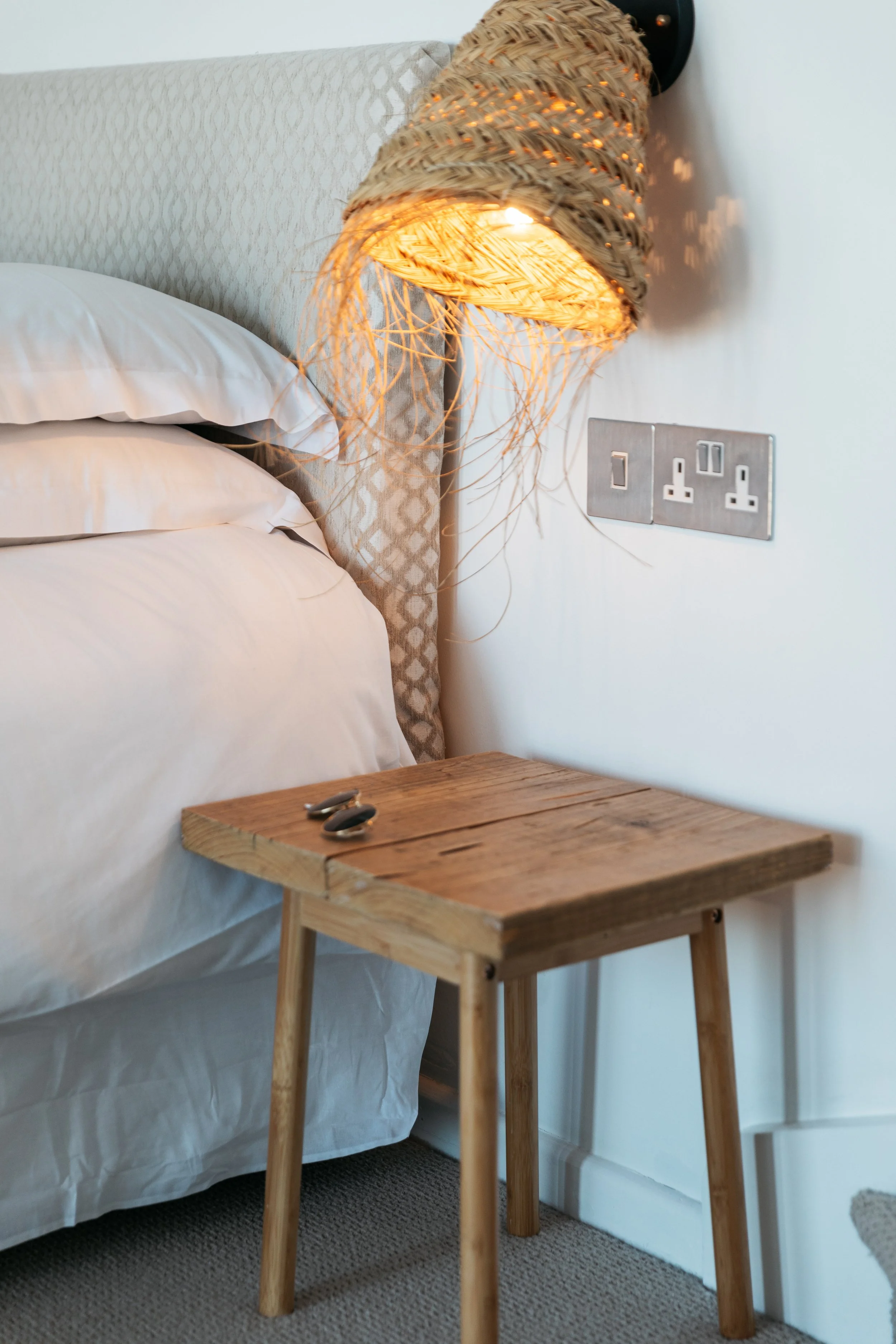 Wooden nightstand next to a bed with a beige headboard and white bedding, beside a wall socket and a woven wall lamp with warm light.