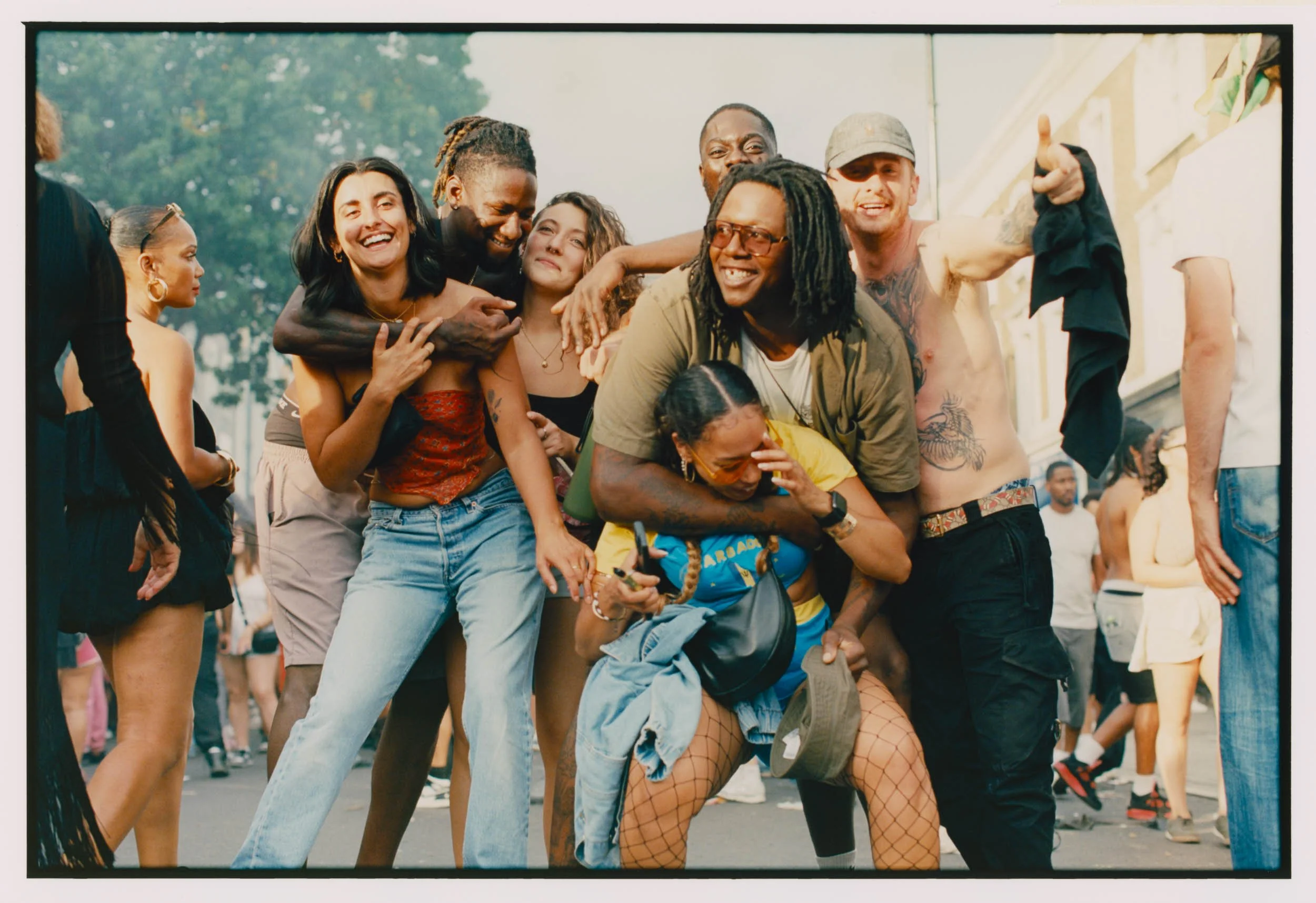 A group of diverse young people posing outdoors during a festival or street event, smiling and interacting with each other, with some people in the background.