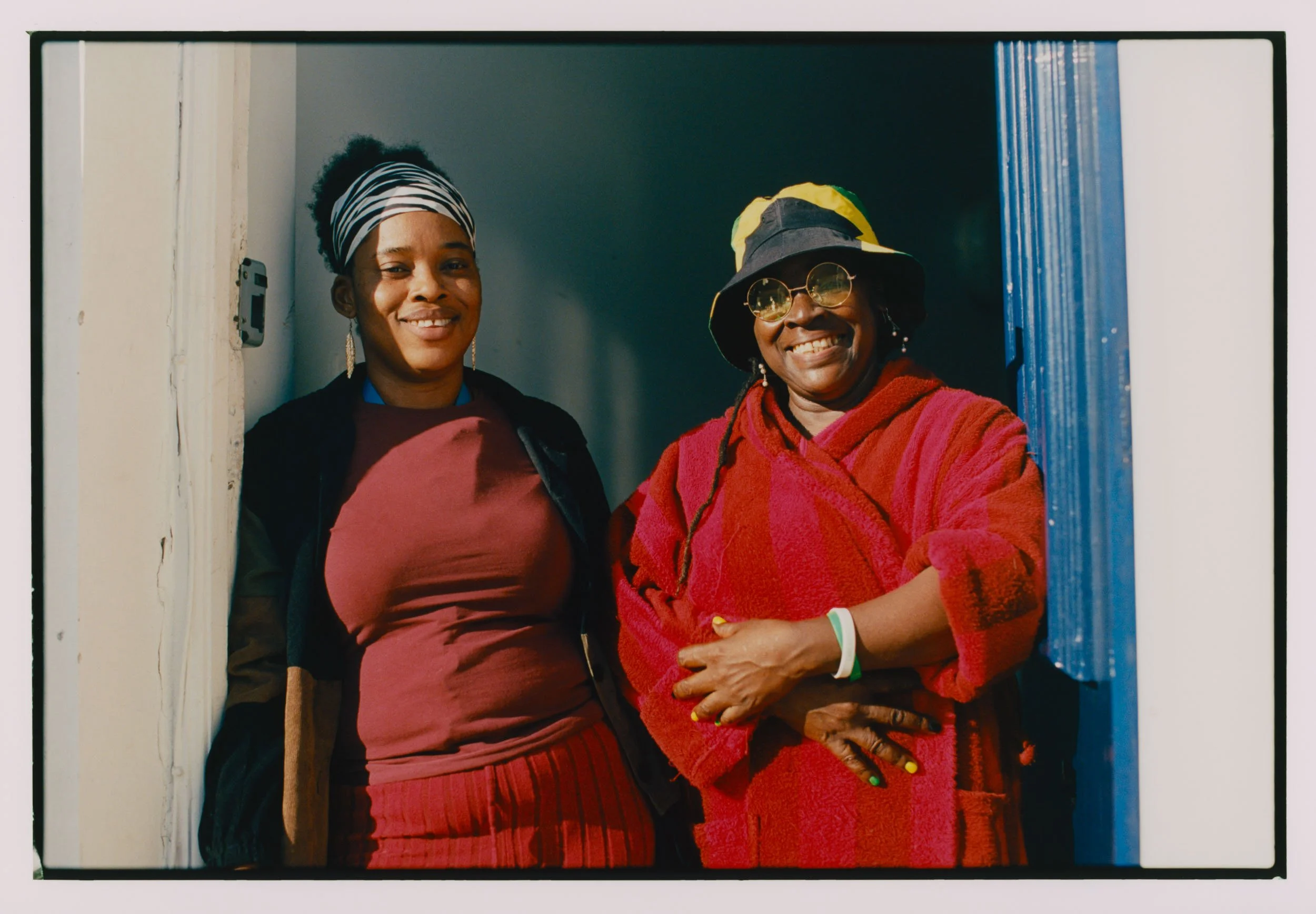 Two women standing together, smiling, wearing colorful and patterned clothing, jewelry, and hats, standing in a doorway.