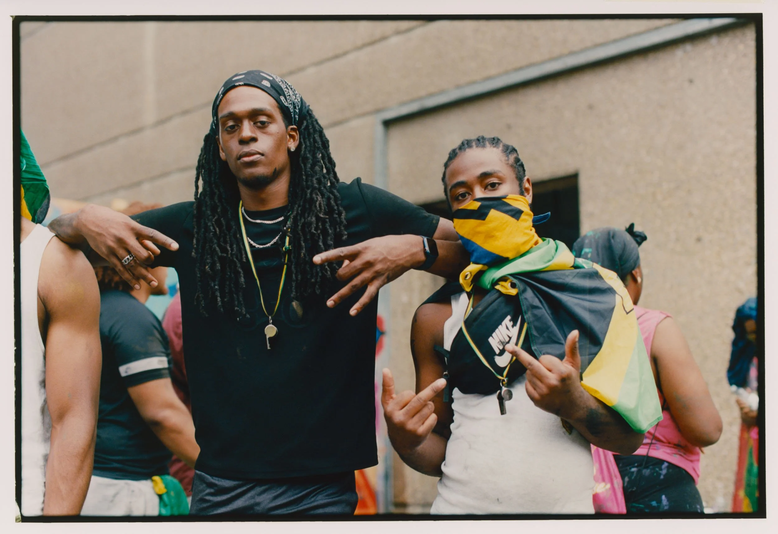 Two young men standing outdoors, one with long dreadlocks and wearing a black t-shirt, the other wearing a yellow bandana mask and a yellow, black, and green jacket, both making hand gestures.
