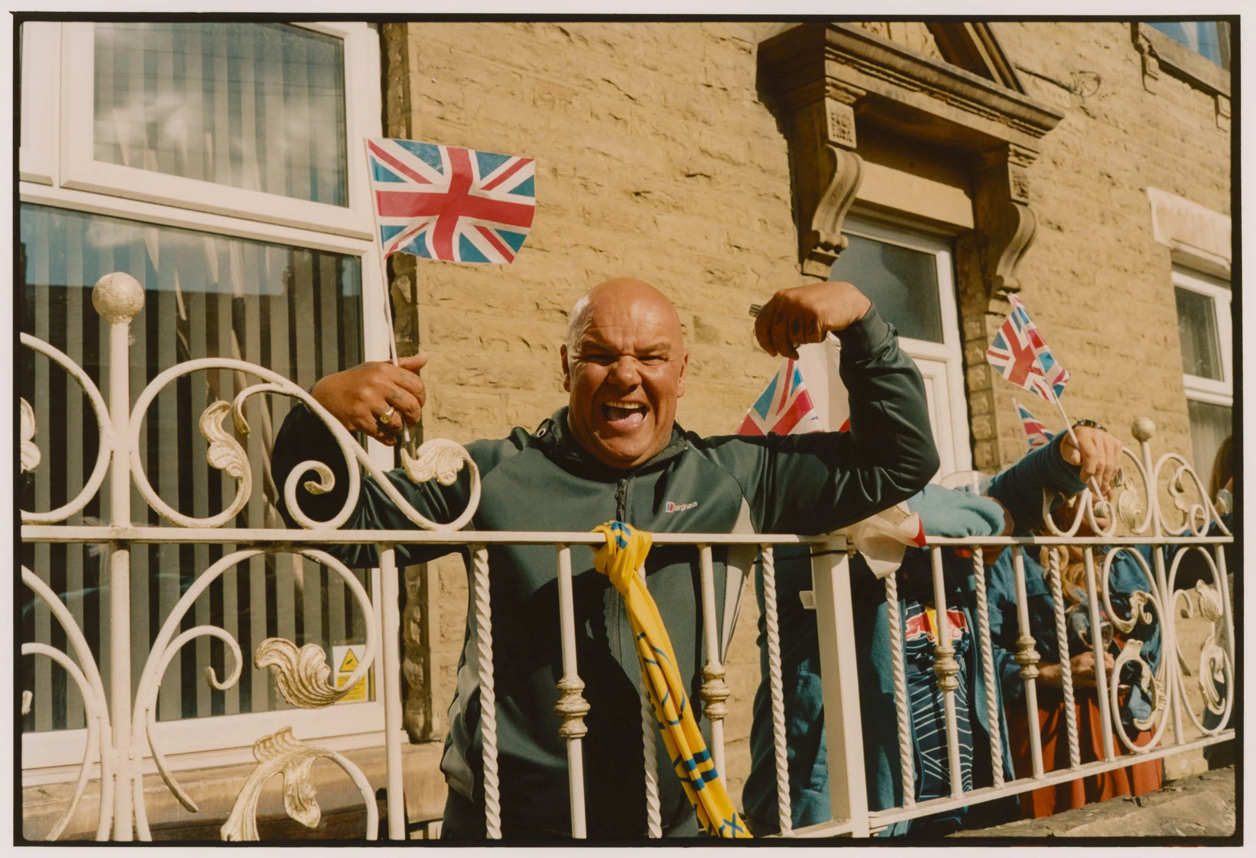 A man with a bald head and gray/silver jacket making a fist and shouting in excitement, standing behind a decorative white metal fence with people and small Union Jack flags visible in the background on a sunny day.
