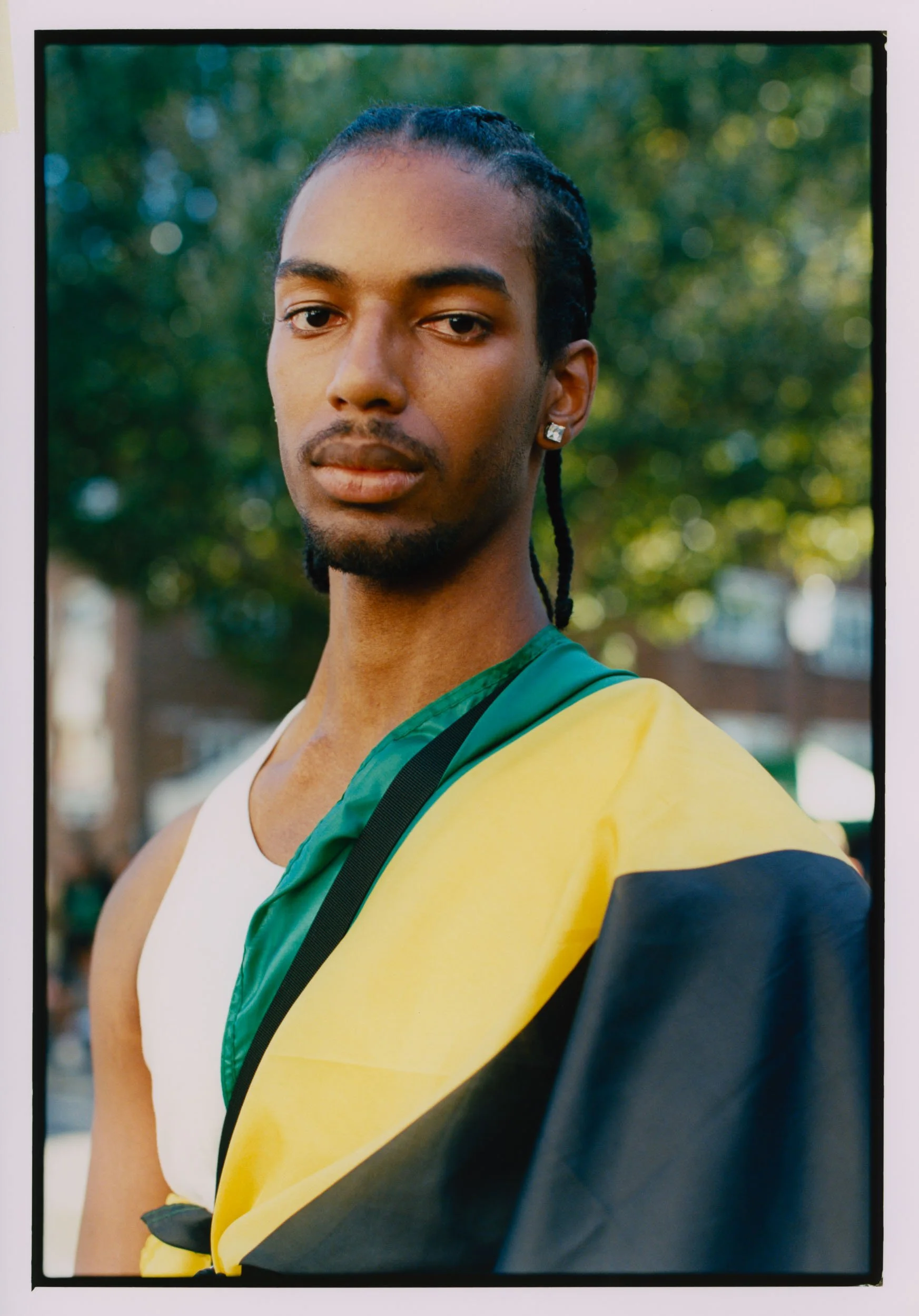 Close-up of a young Black man with braided hair, wearing a white tank top and yellow and black jacket, sitting on a black chair outdoors with a blurred background of green trees and sunlight.