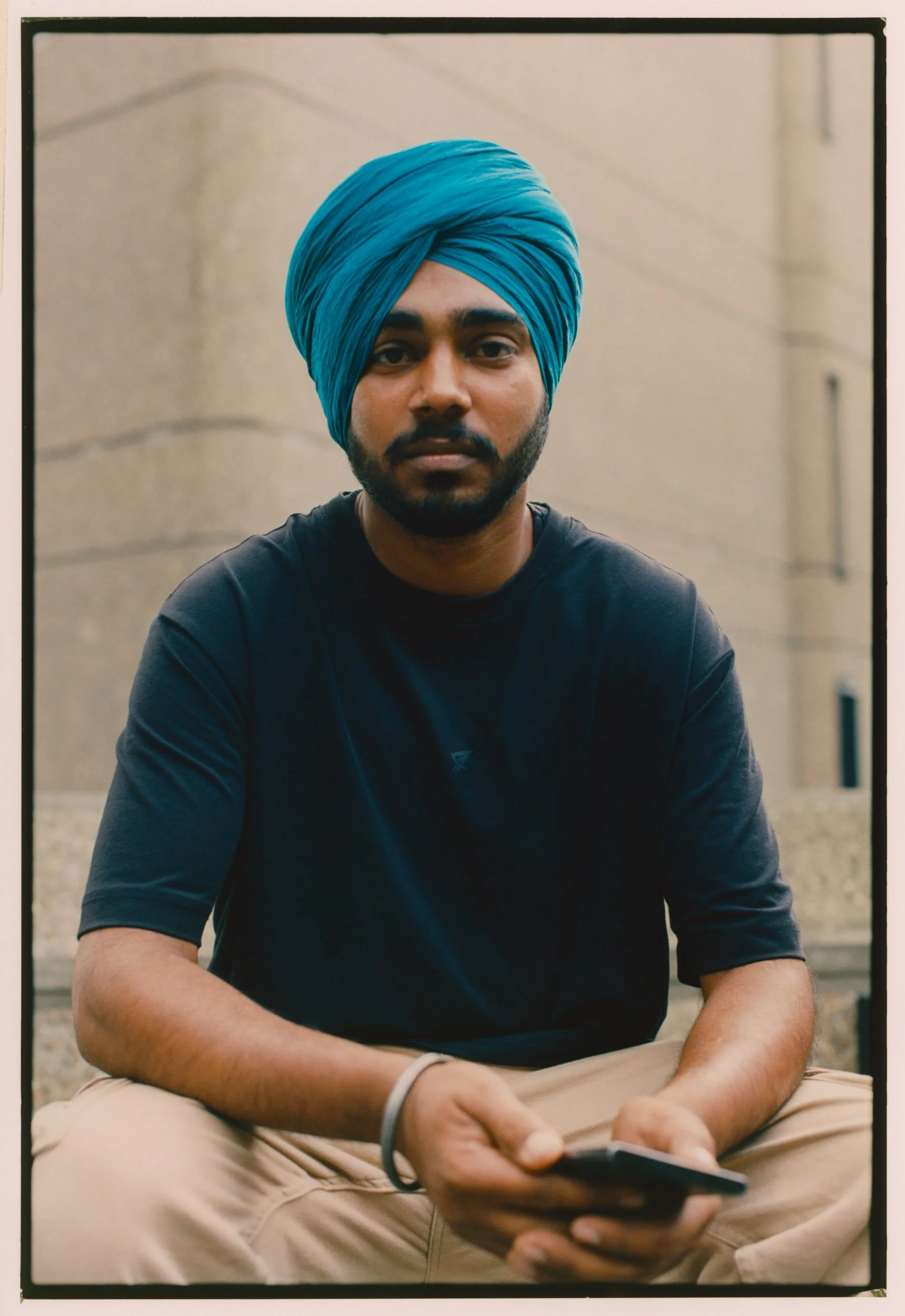 A young man wearing a blue turban and black T-shirt seated outside on a concrete surface, holding a smartphone in his hand.