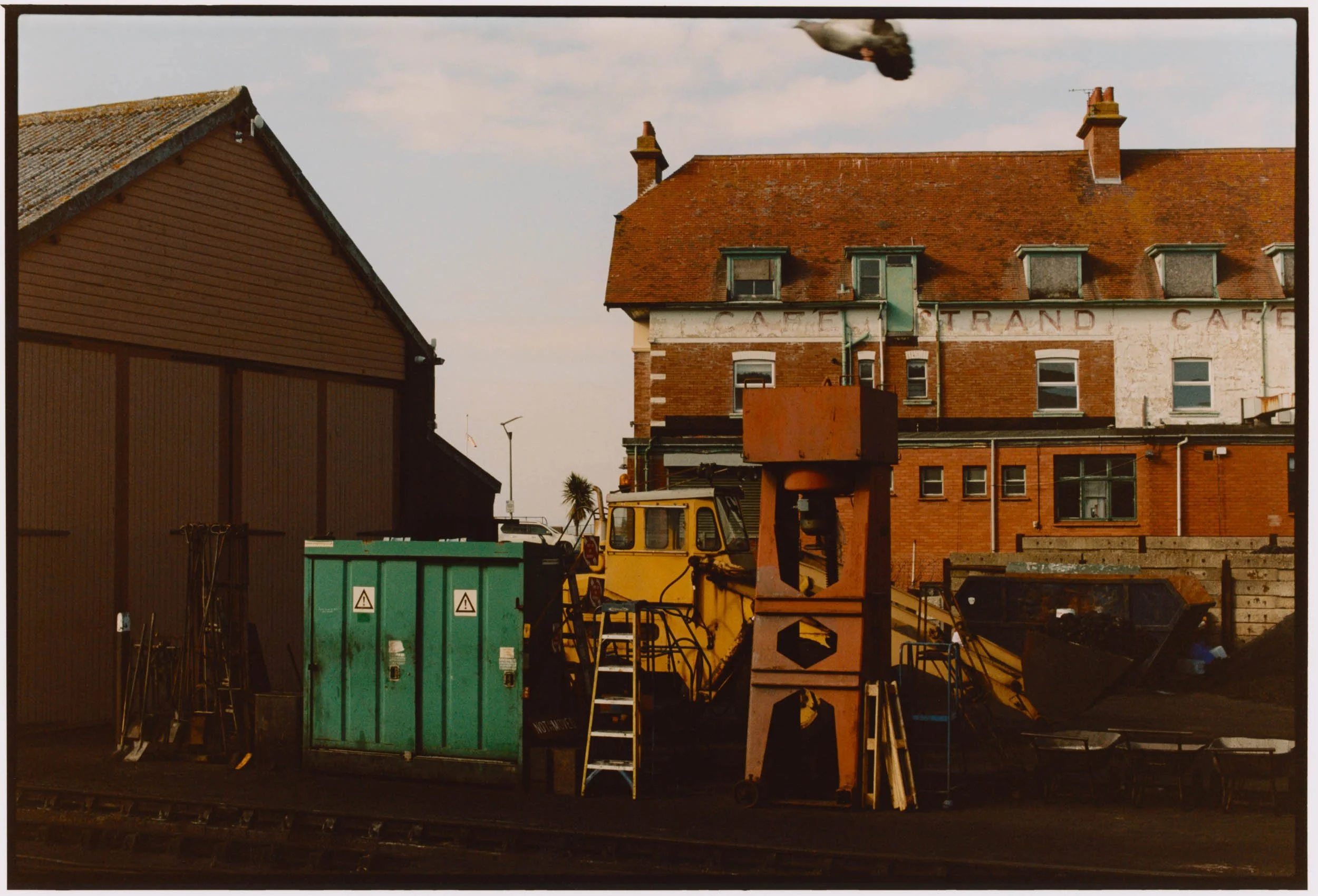 An urban scene showing a brick building with a red tiled roof, a pigeon flying overhead, construction equipment including a yellow vehicle, green containers, and various construction materials in the foreground.