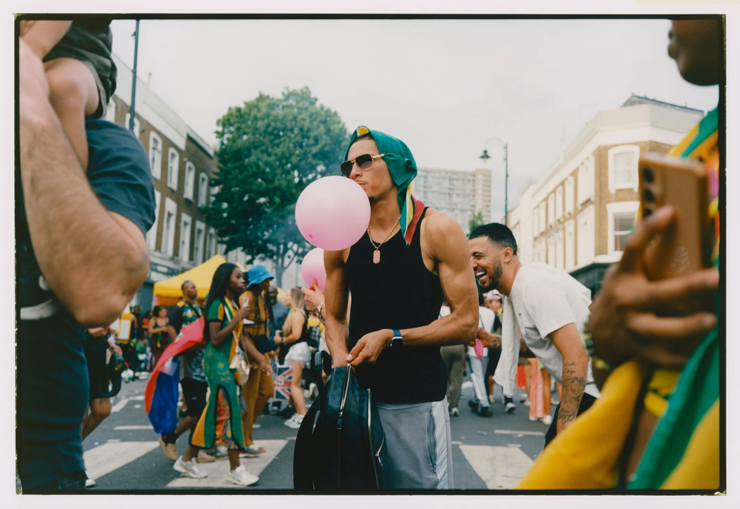 Group of people celebrating at an outdoor street event, some wearing rainbow colors and carrying balloons, with buildings and trees in the background.