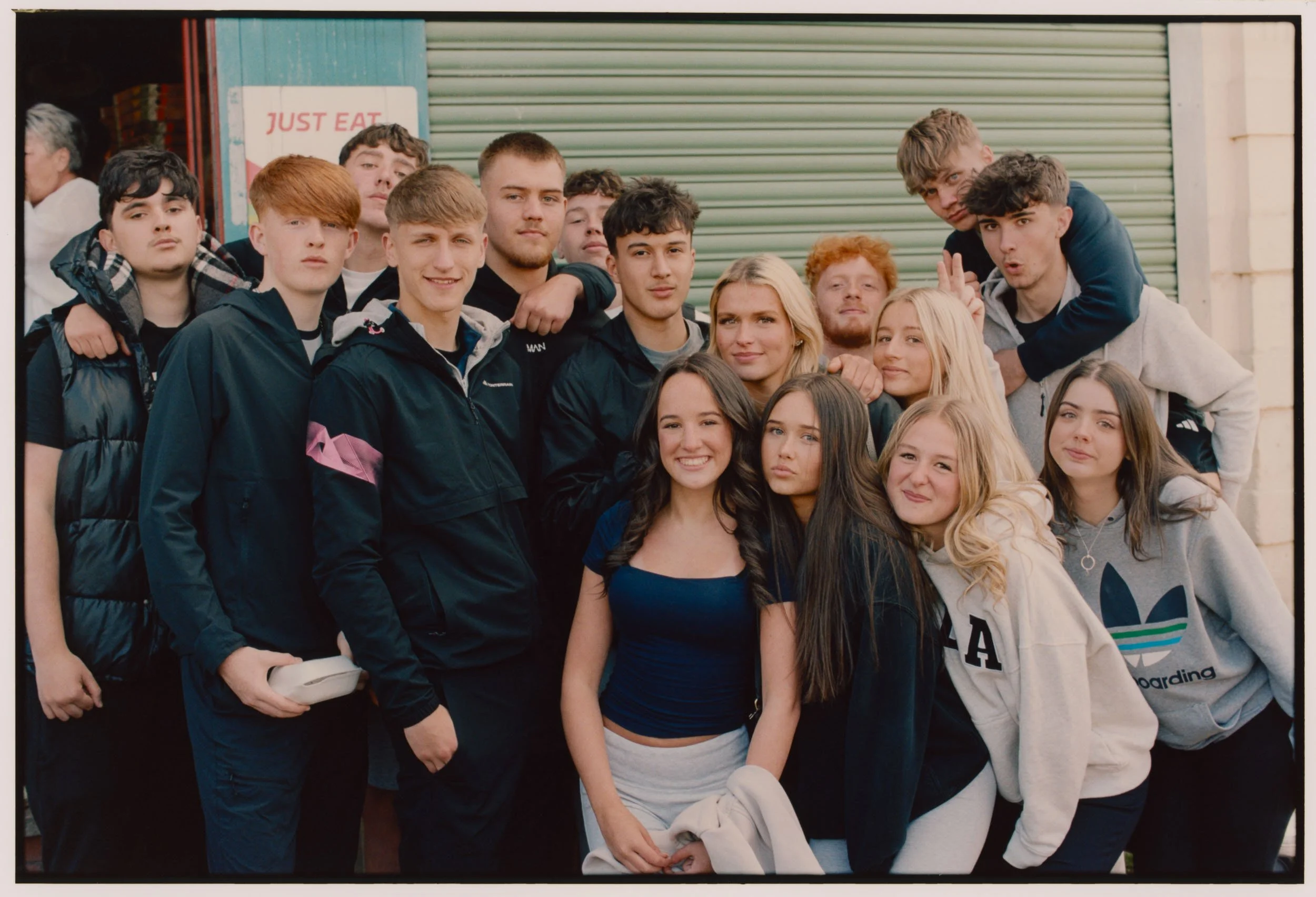 A group of young people, mostly males, posing together outdoors in front of a green roll-up door and a wall, with some making peace signs, smiling and looking at the camera.