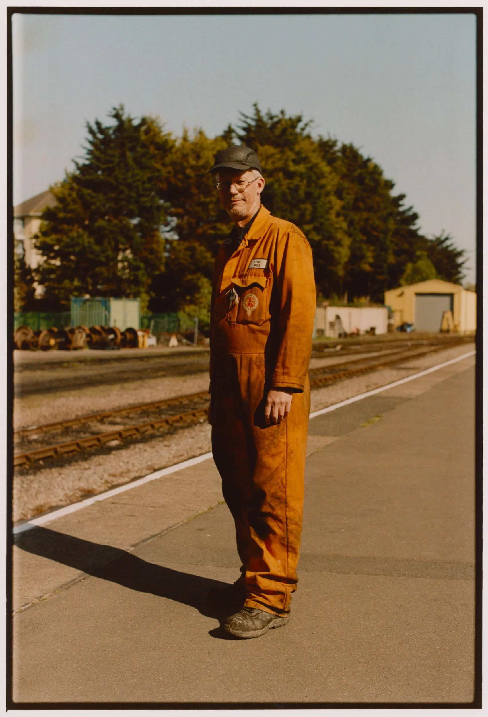 A man in an orange jumpsuit standing on a concrete platform beside railroad tracks, with trees and small buildings in the background, during daytime.