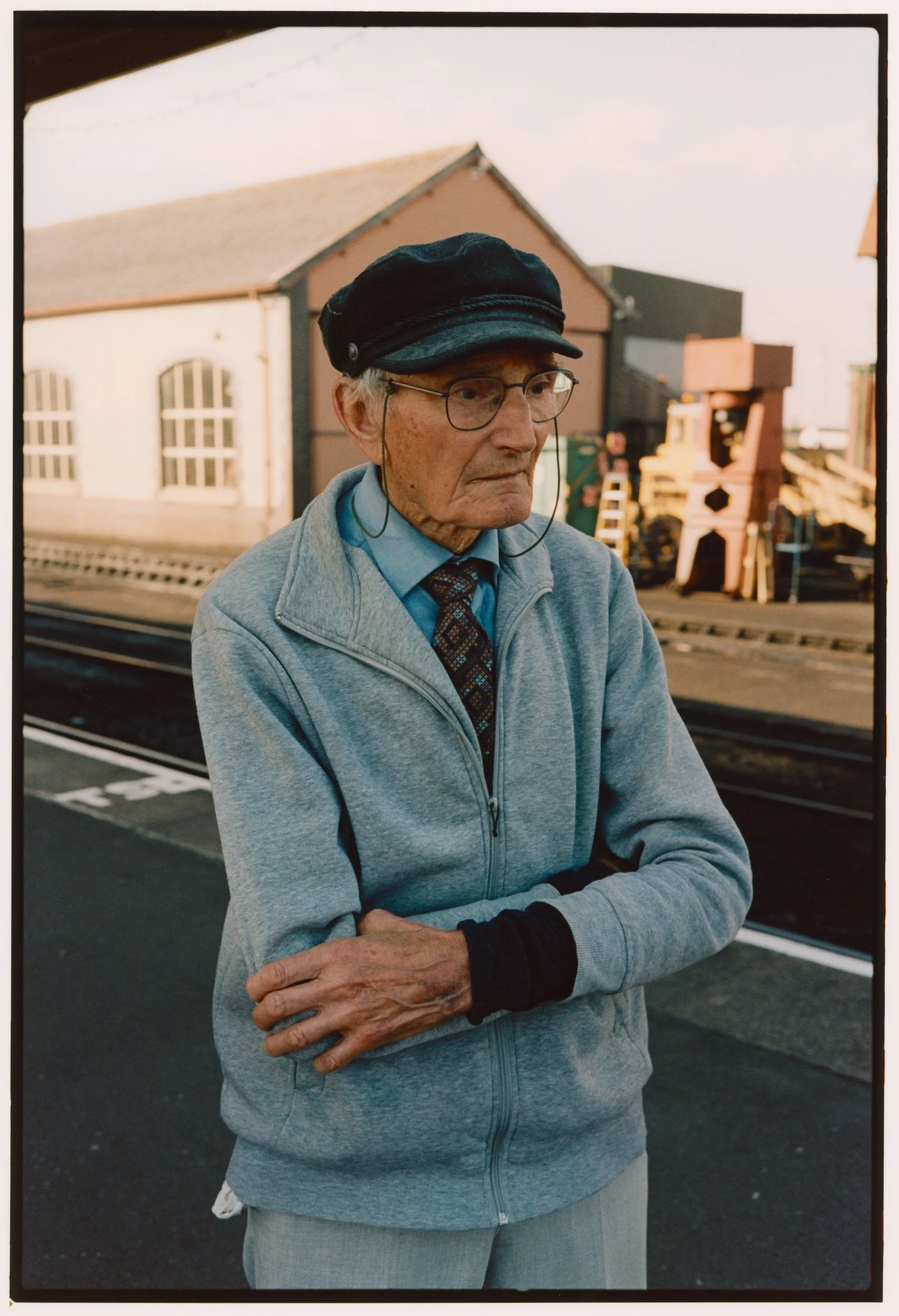 An elderly man with glasses, a black cap, and a gray zip-up hoodie stands outdoors with arms crossed. Behind him are train tracks and a warehouse-like building.