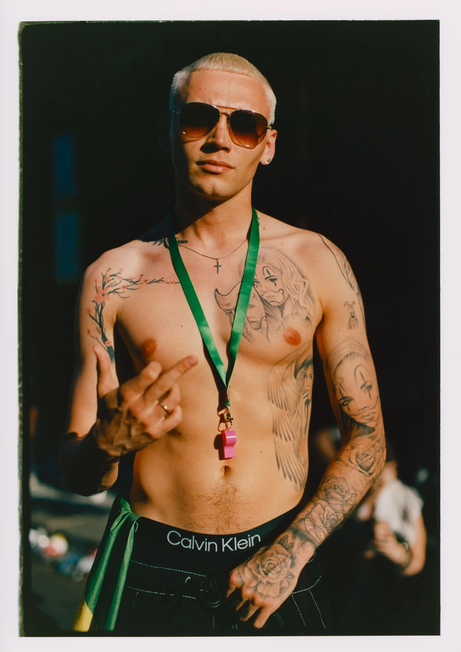 A shirtless man wearing sunglasses, a cross necklace, Adidas pants, and a green lanyard, with tattoos on his chest and arms, standing outdoors with a blurred background.