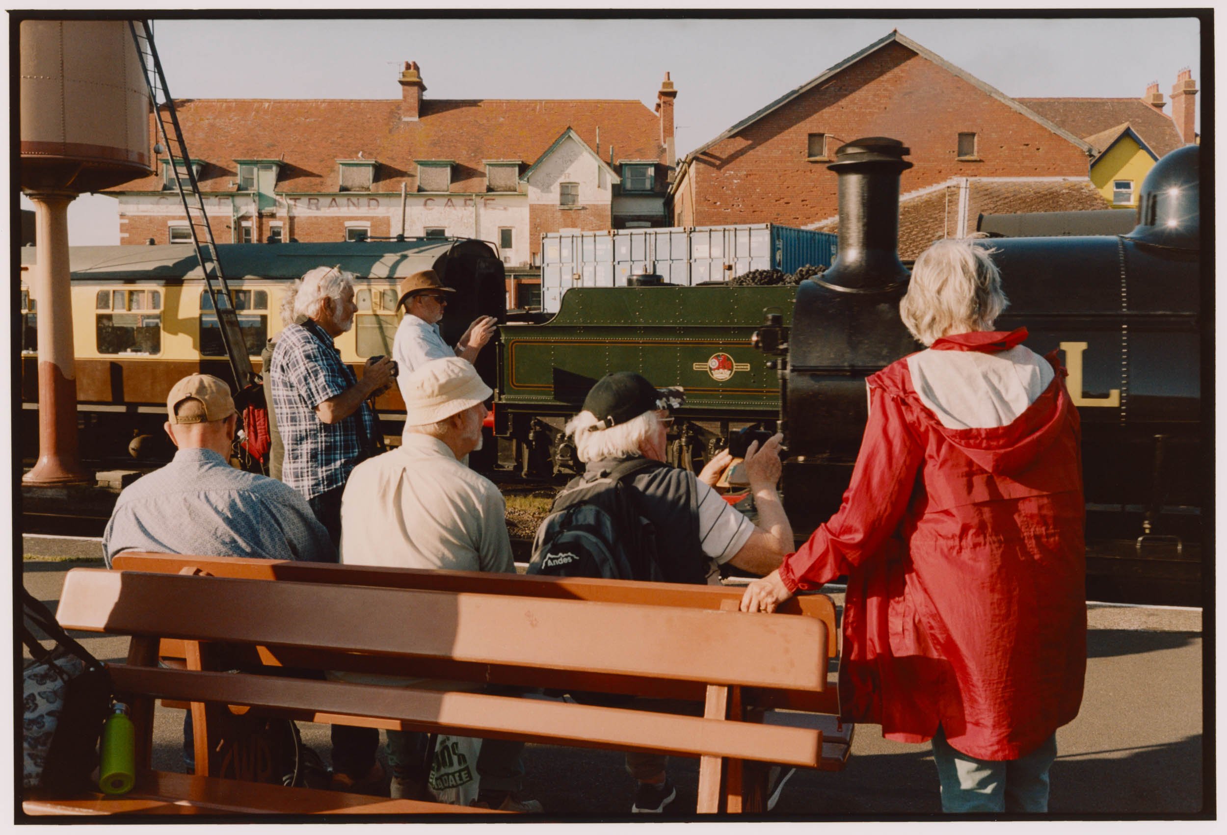 Group of people gathered outdoors around a vintage steam locomotive train, some sitting on benches, others standing and taking photos. Houses with brick walls and chimneys are visible in the background.