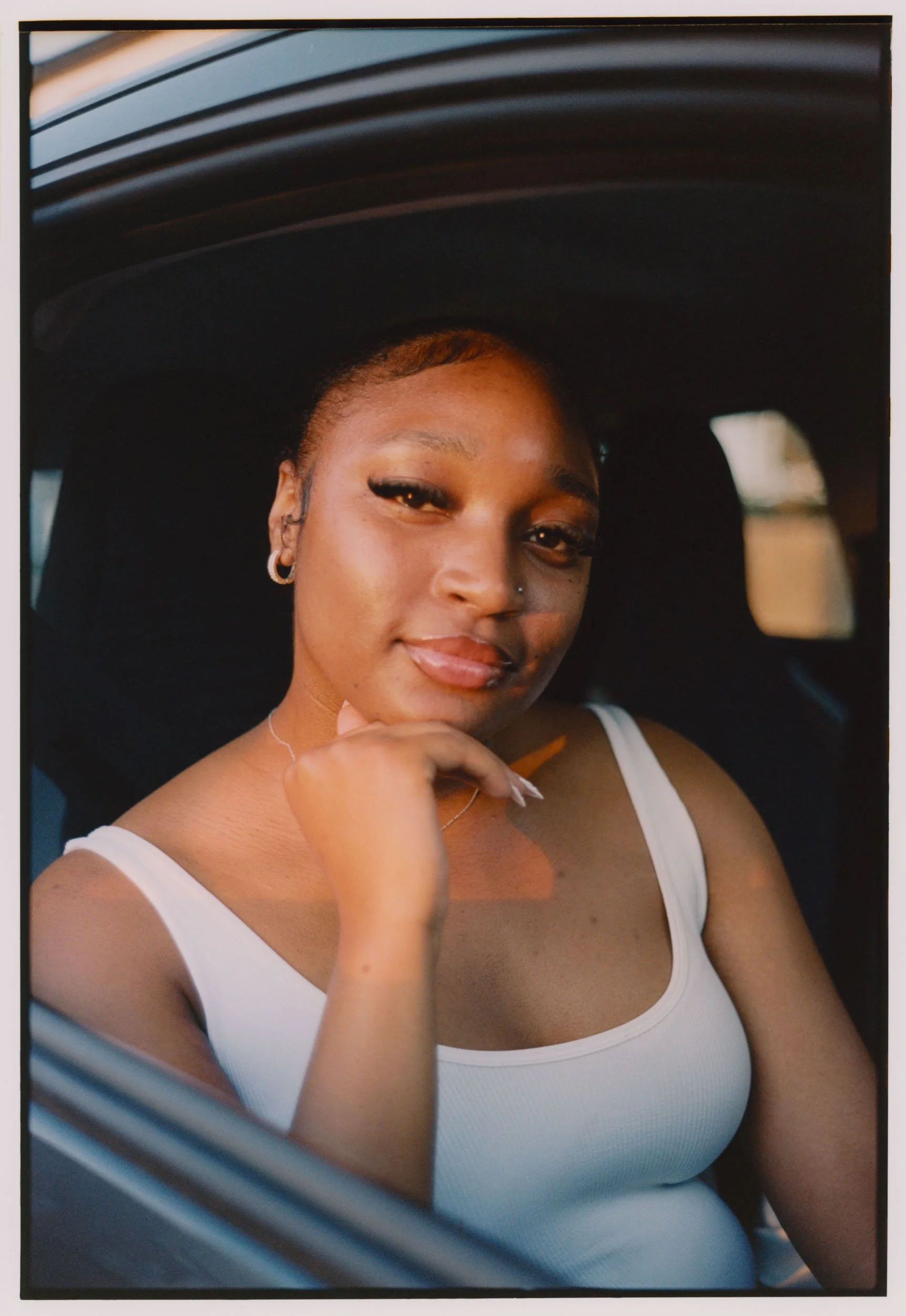 A young woman with short hair, dark skin, and hoop earrings sitting in a car, smiling at the camera, wearing a white tank top.