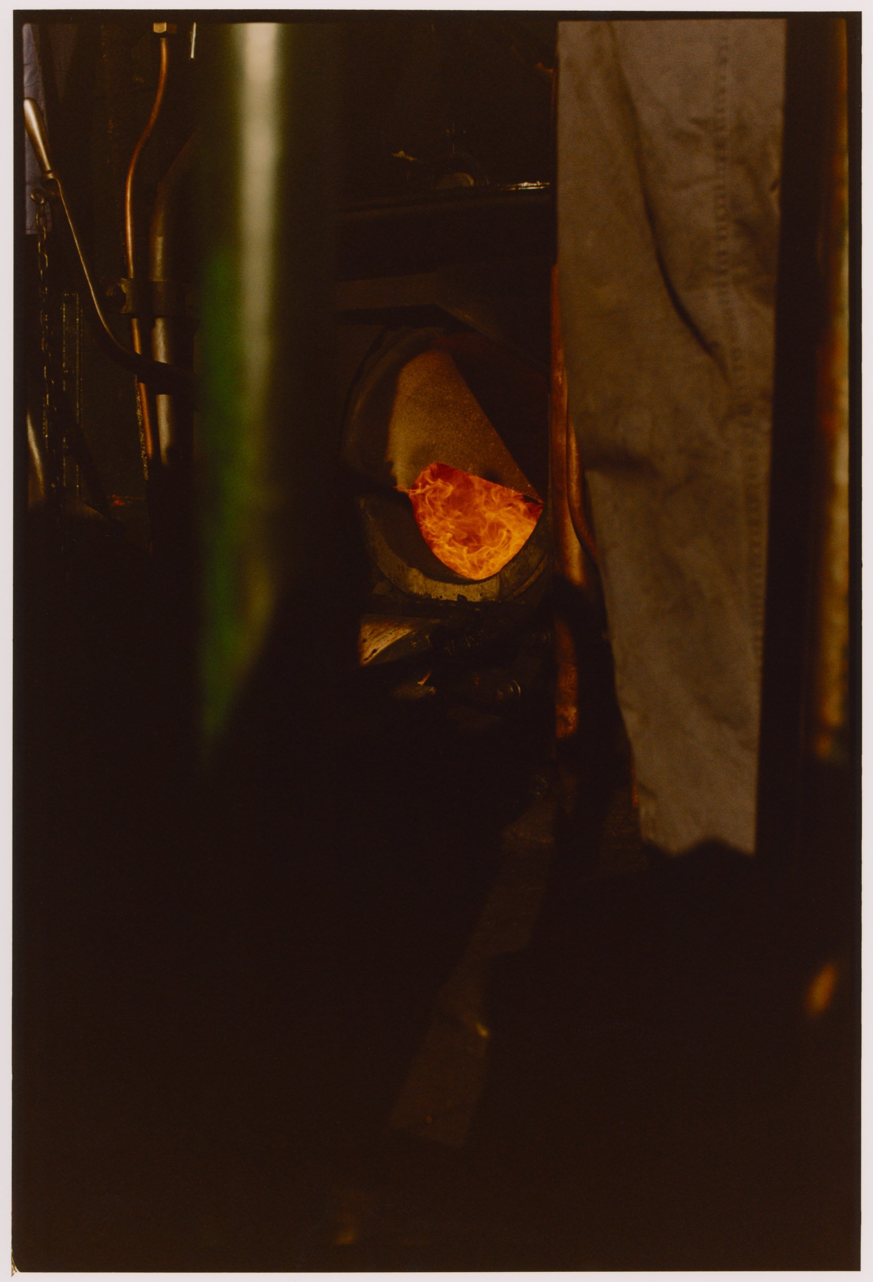 A blurry image with a dark environment. There is a glowing, fiery object or light source in the center, possibly resembling a rock or molten material. Some metallic or mechanical parts are visible around the edges.