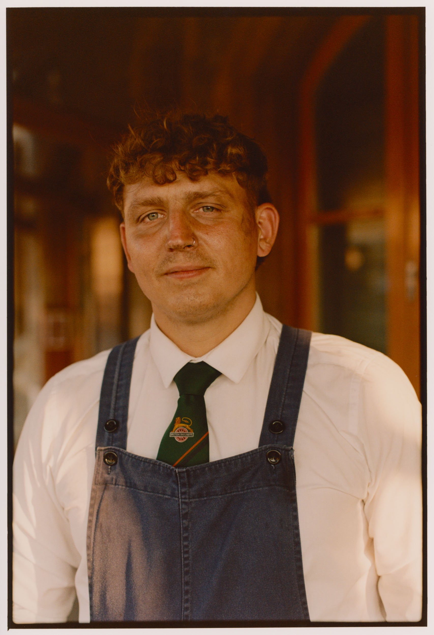 A man with curly hair wearing a white shirt, black tie, and denim apron, standing indoors with a warm wooden background.
