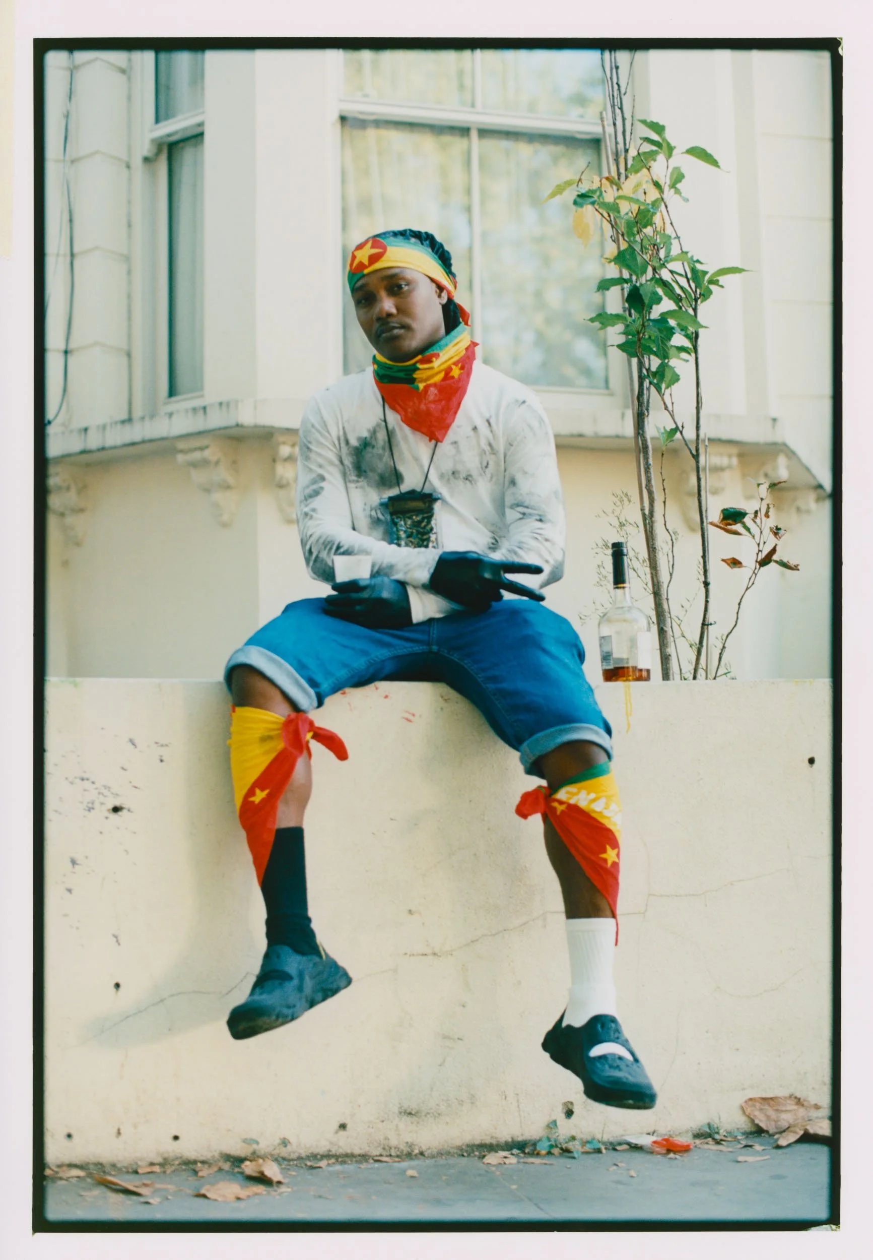A person sitting on a ledge in front of a wall and window, wearing a multicolored headband and neck warmer, a white distressed long-sleeve shirt, blue shorts, socks, and sneakers, with a bottle of alcohol on the ground beside them and leaves on the g