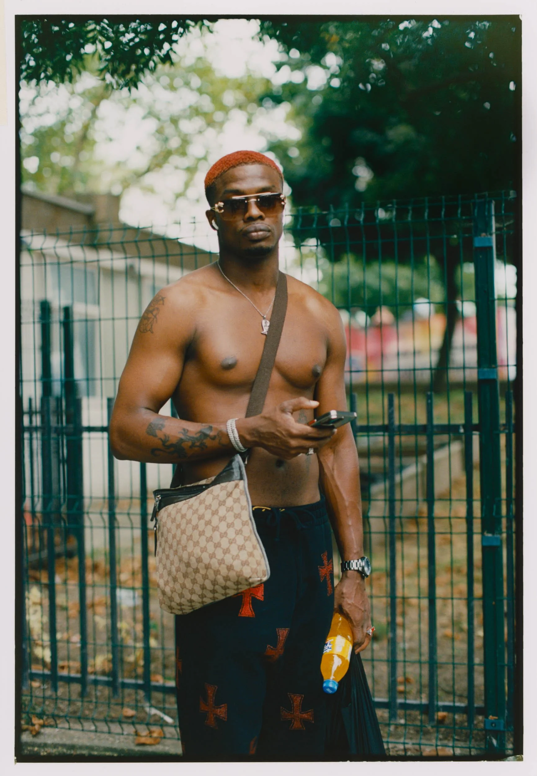 A topless man with sunglasses and red hair, holding a phone, standing outdoors near a fence, with trees in the background.