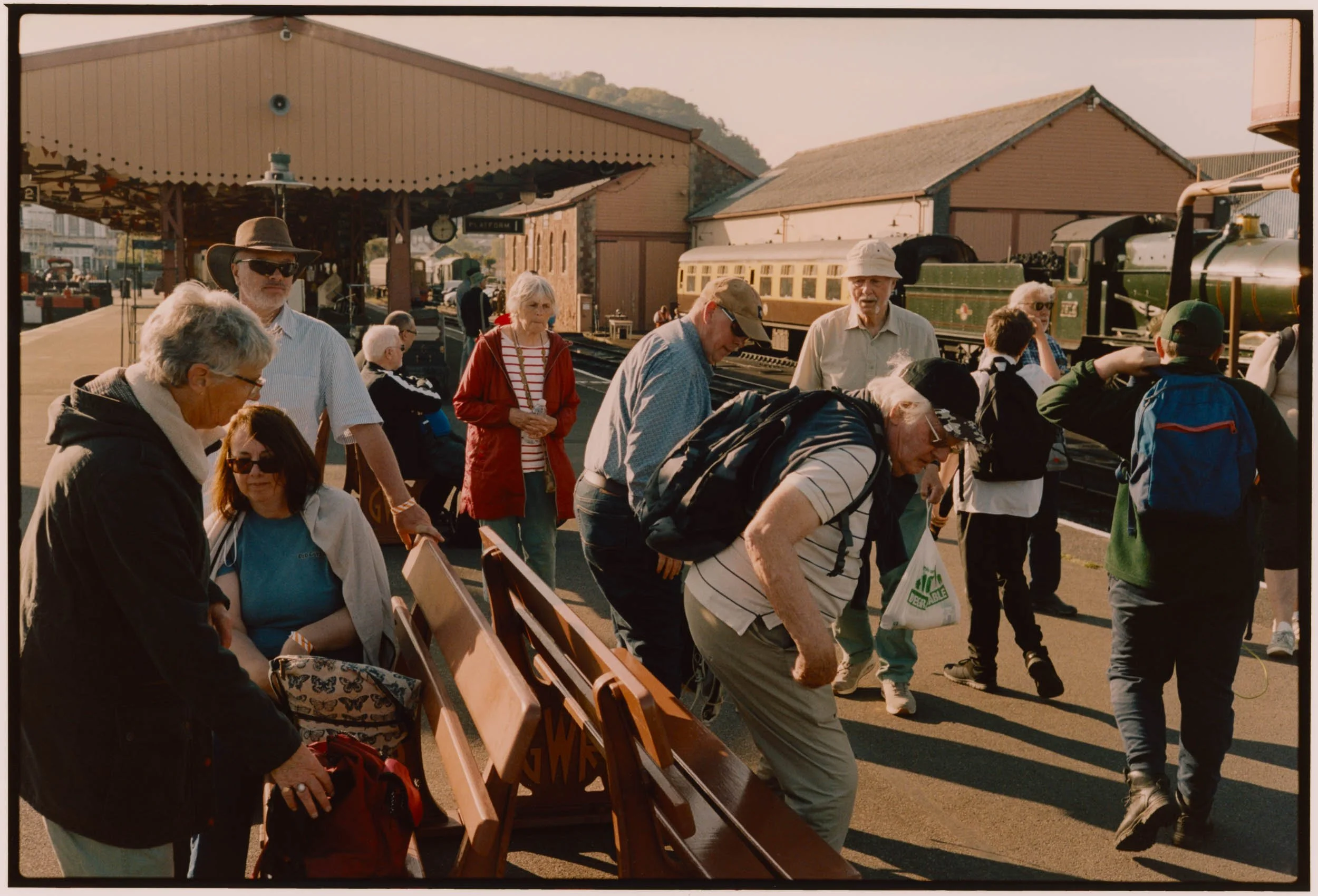 A crowd of people gathered near train tracks, with a vintage green steam locomotive visible in the background on the right side of the image. Some individuals are standing, sitting, or looking at the train, and several are wearing hats and sunglasses