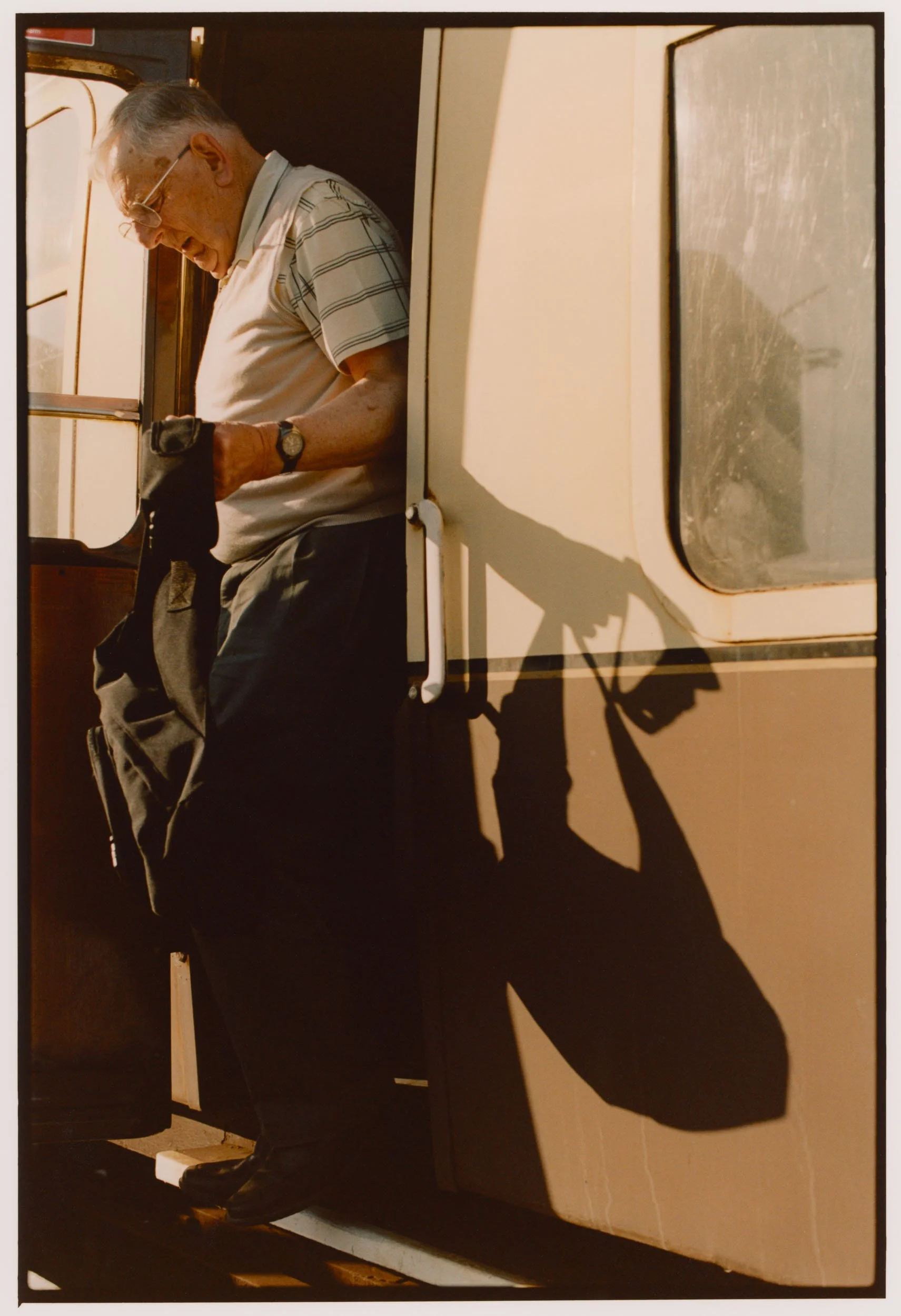 An elderly man with gray hair and glasses, wearing a striped shirt, black pants, and a watch, standing in front of a door with a window, holding a black bag and looking down.