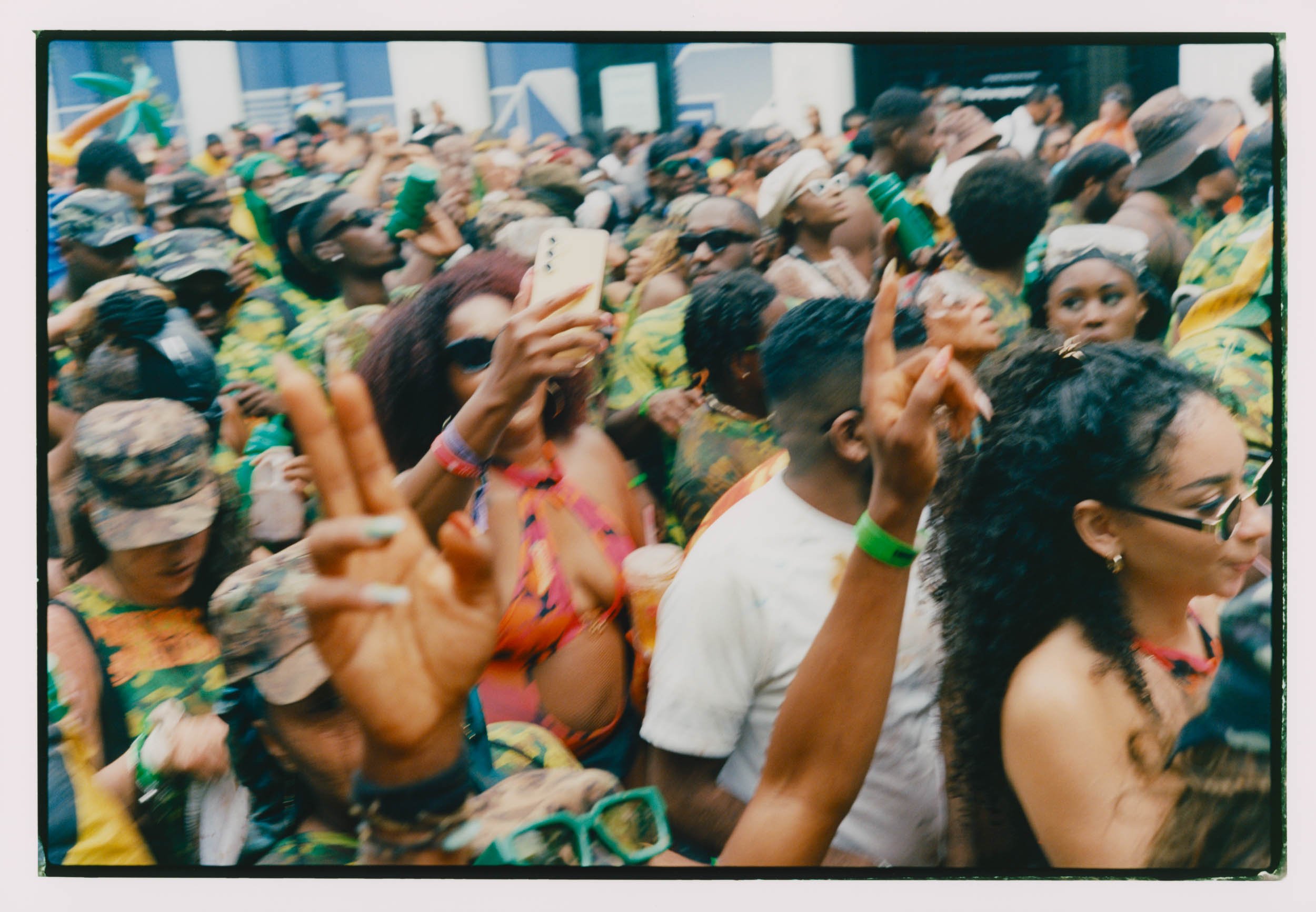 A large crowd of people gathering outdoors during a festive event, with some holding up phones and wearing colorful clothing and accessories.
