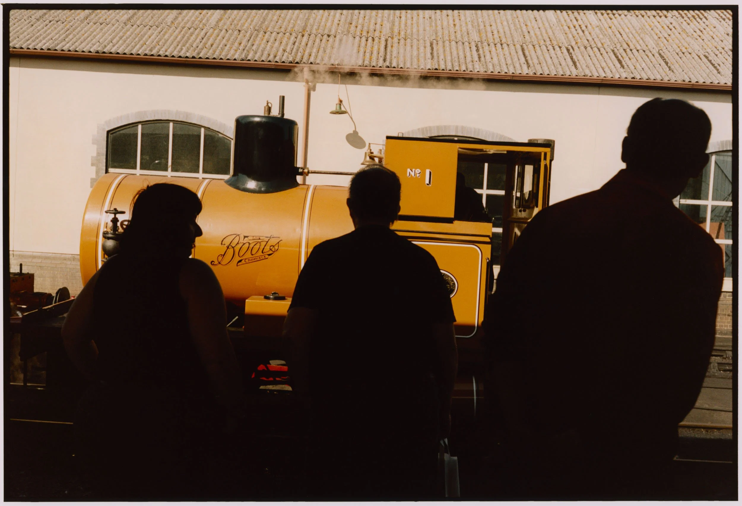 People standing in front of a yellow steam engine train with a black chimney, small windows, and the word 'Boots' on the side, outside a building with a tiled roof and large window.