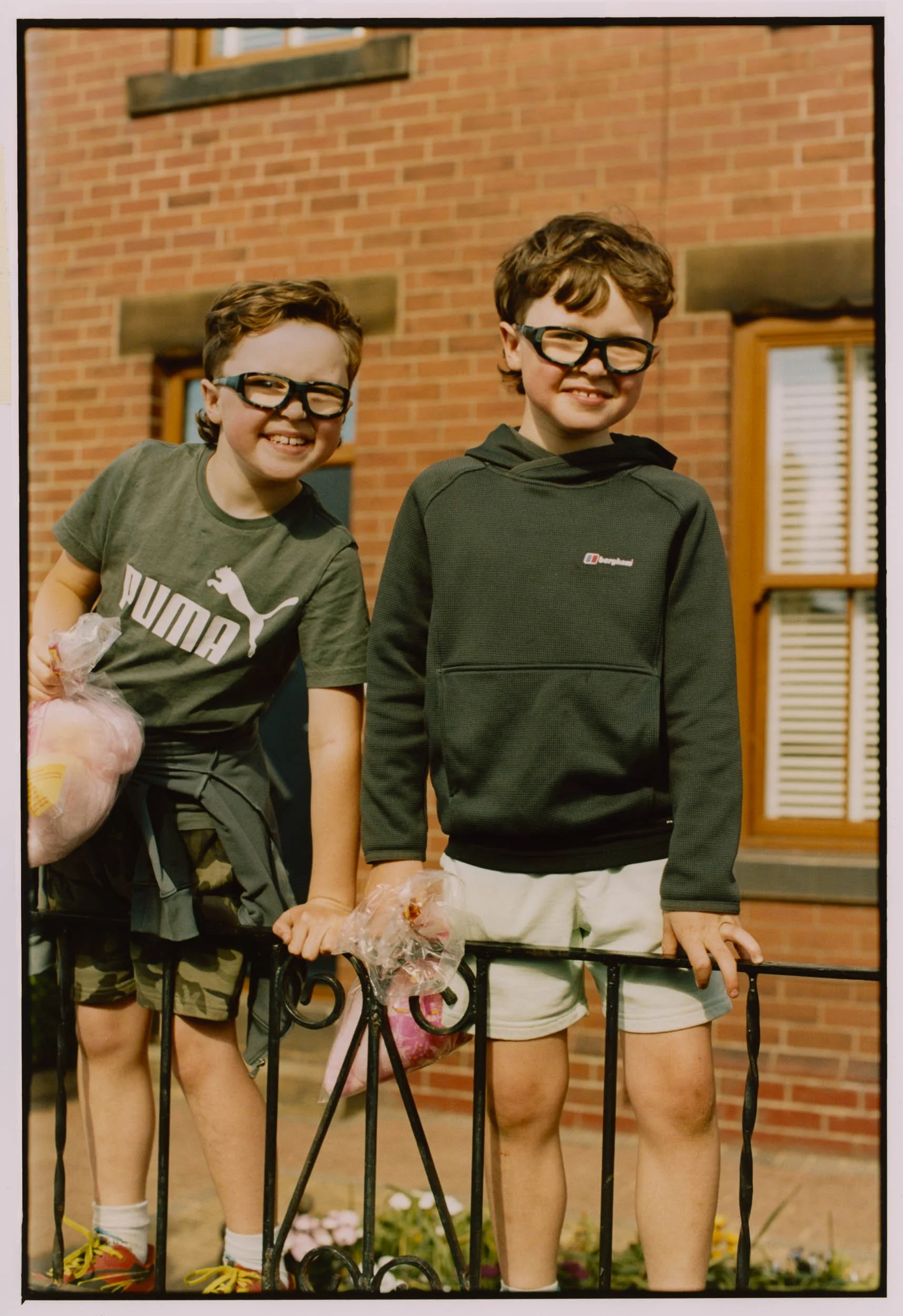 Two young boys smiling and holding hands, standing behind a black iron fence in front of a brick building, each carrying a bag of cotton candy.