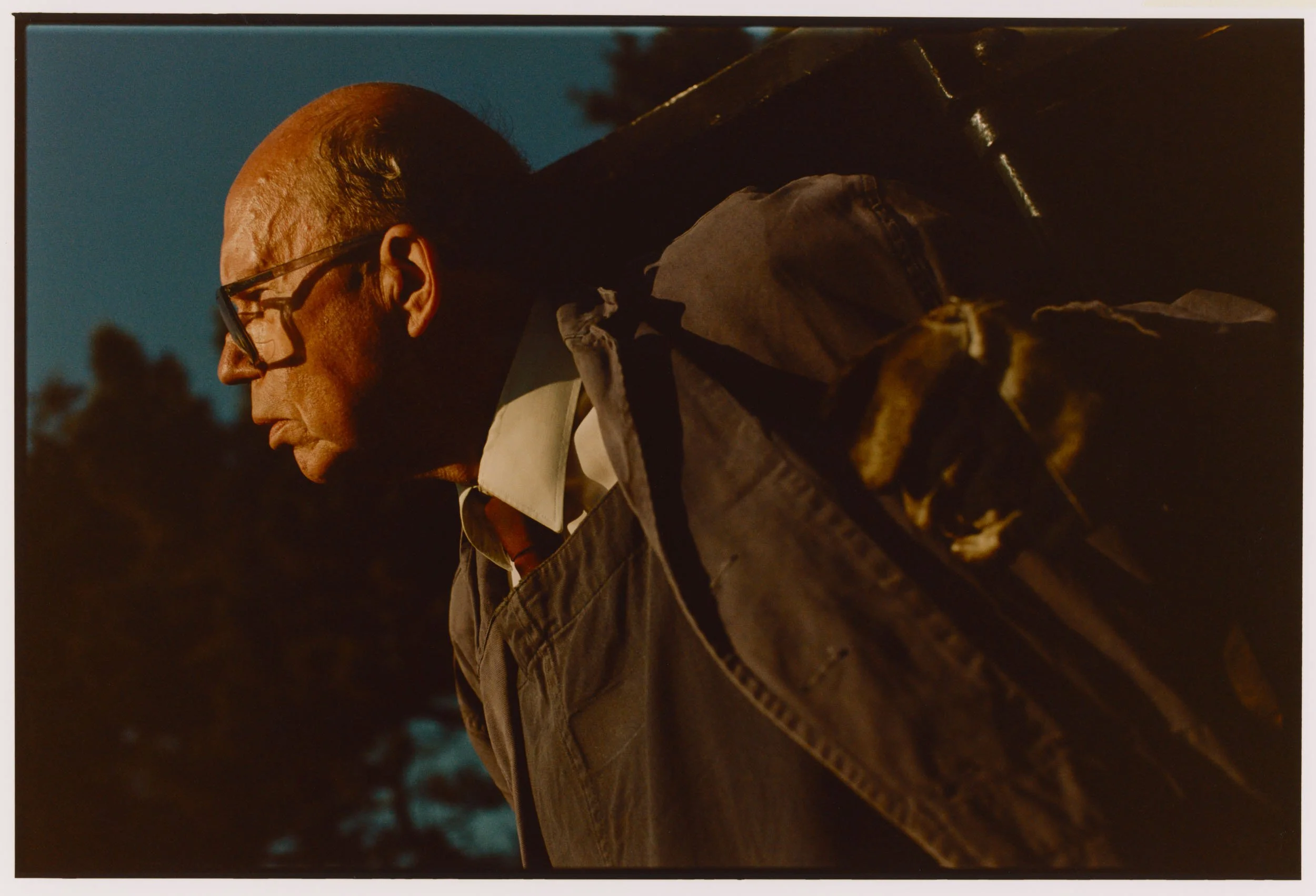 A man with glasses in profile view, wearing a jacket, standing outdoors during night or evening with a dark sky and trees in the background.