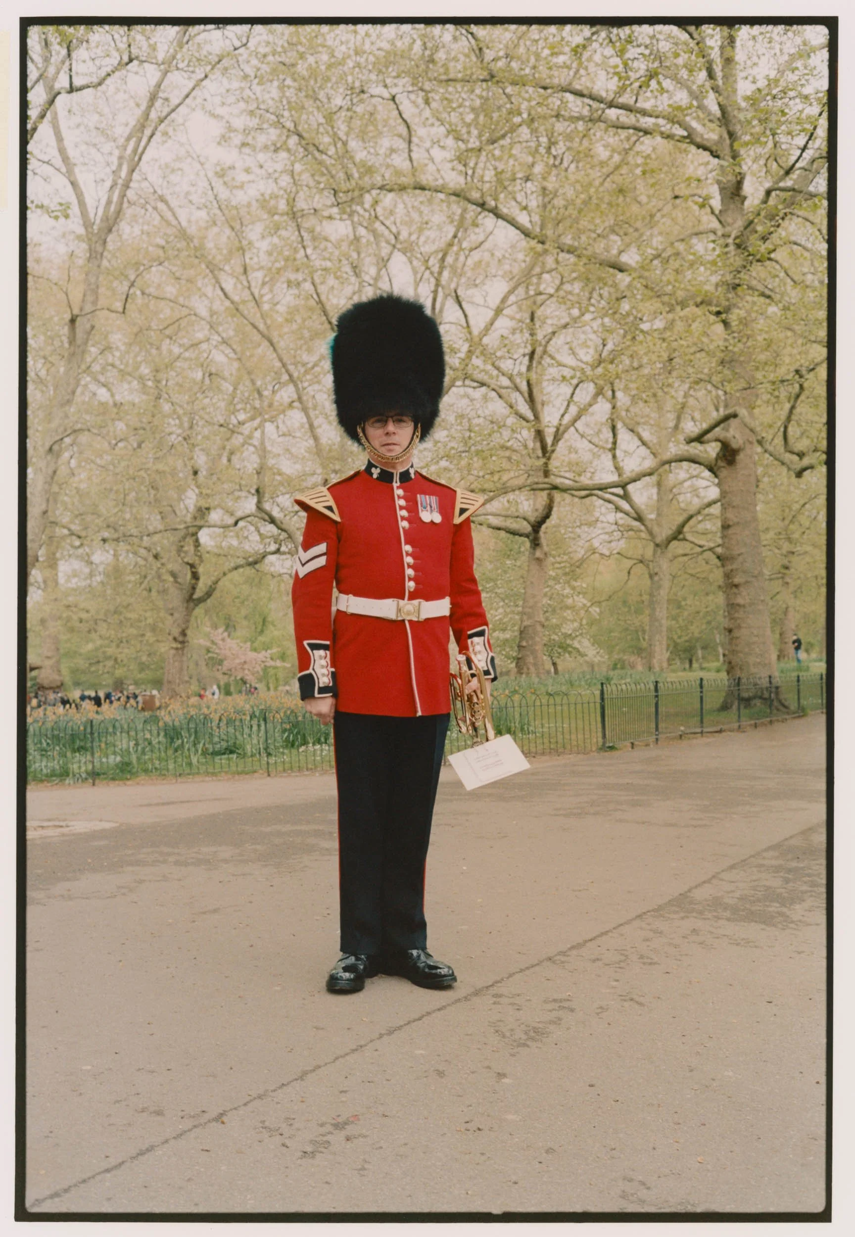 A soldier in a red dress uniform with a tall black bearskin hat standing outdoors in front of trees with light-colored leaves.