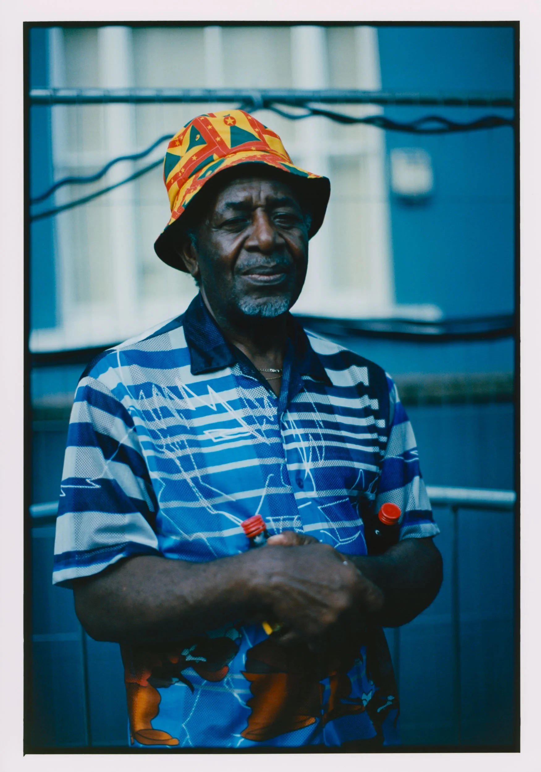 Older man standing with arms crossed, holding two bottles with orange caps, wearing a colorful bucket hat with star patterns, and a blue striped shirt, in front of a metallic background.