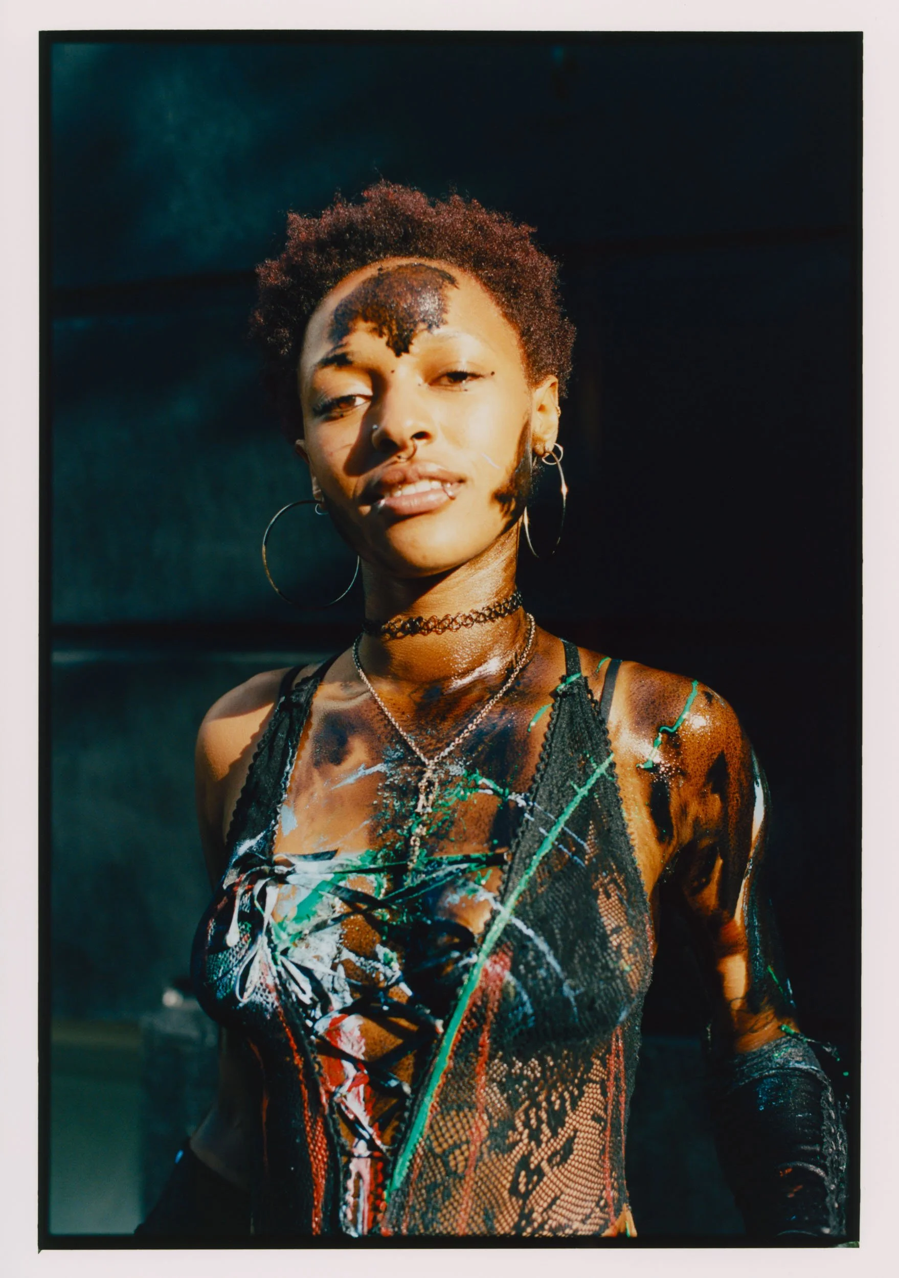 A young woman with short curly hair, wearing hoop earrings, a choker, layered necklaces, and a colorful, abstract-patterned top or dress, posing outdoors in natural light.