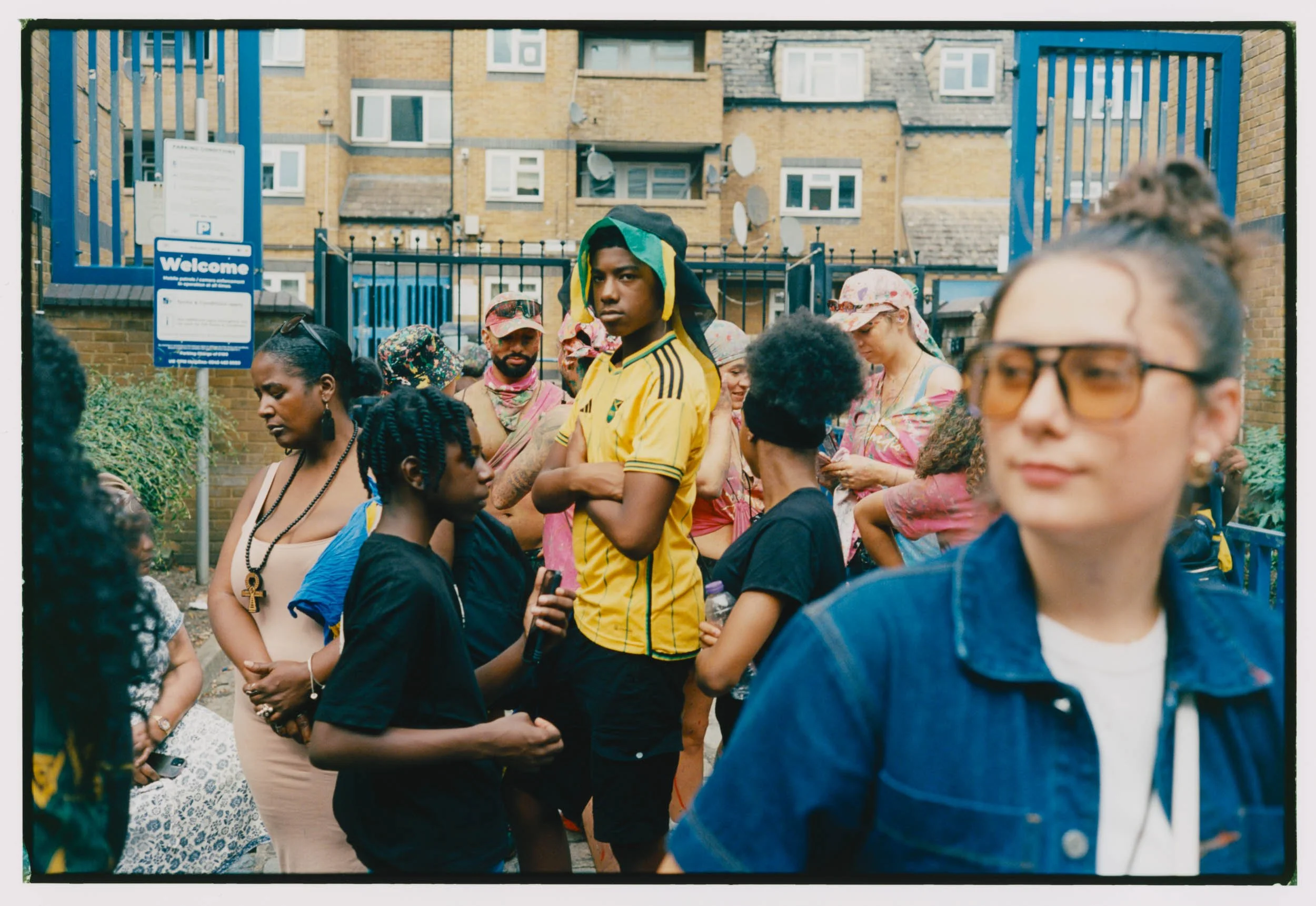 A diverse group of people gathered outdoors near a blue gate and fence, with multi-story brick buildings in the background. The group includes individuals of different ethnicities, some wearing colorful clothing and accessories, others in casual atti