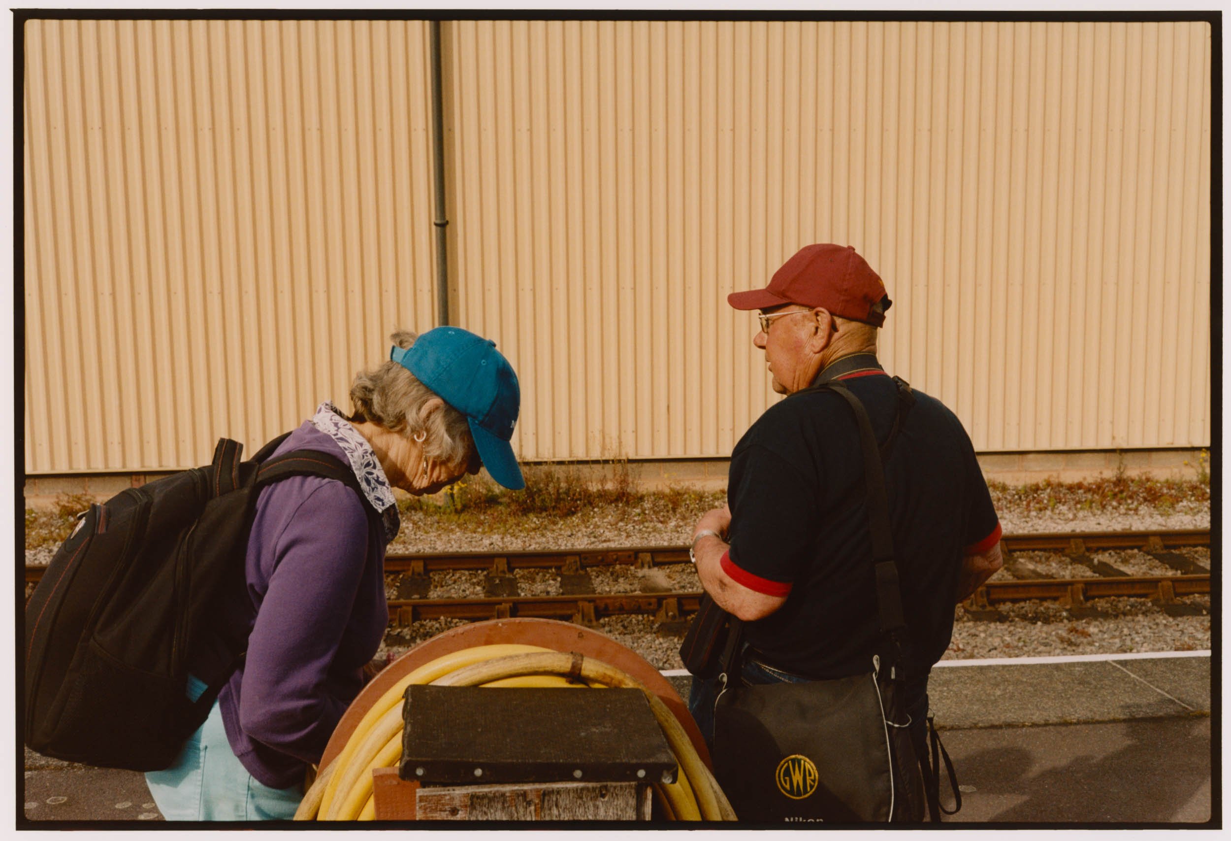 Two elderly people with backpacks, a woman with a blue cap and a man with a red cap, are standing by train tracks looking down at a large pumpkin-shaped container.
