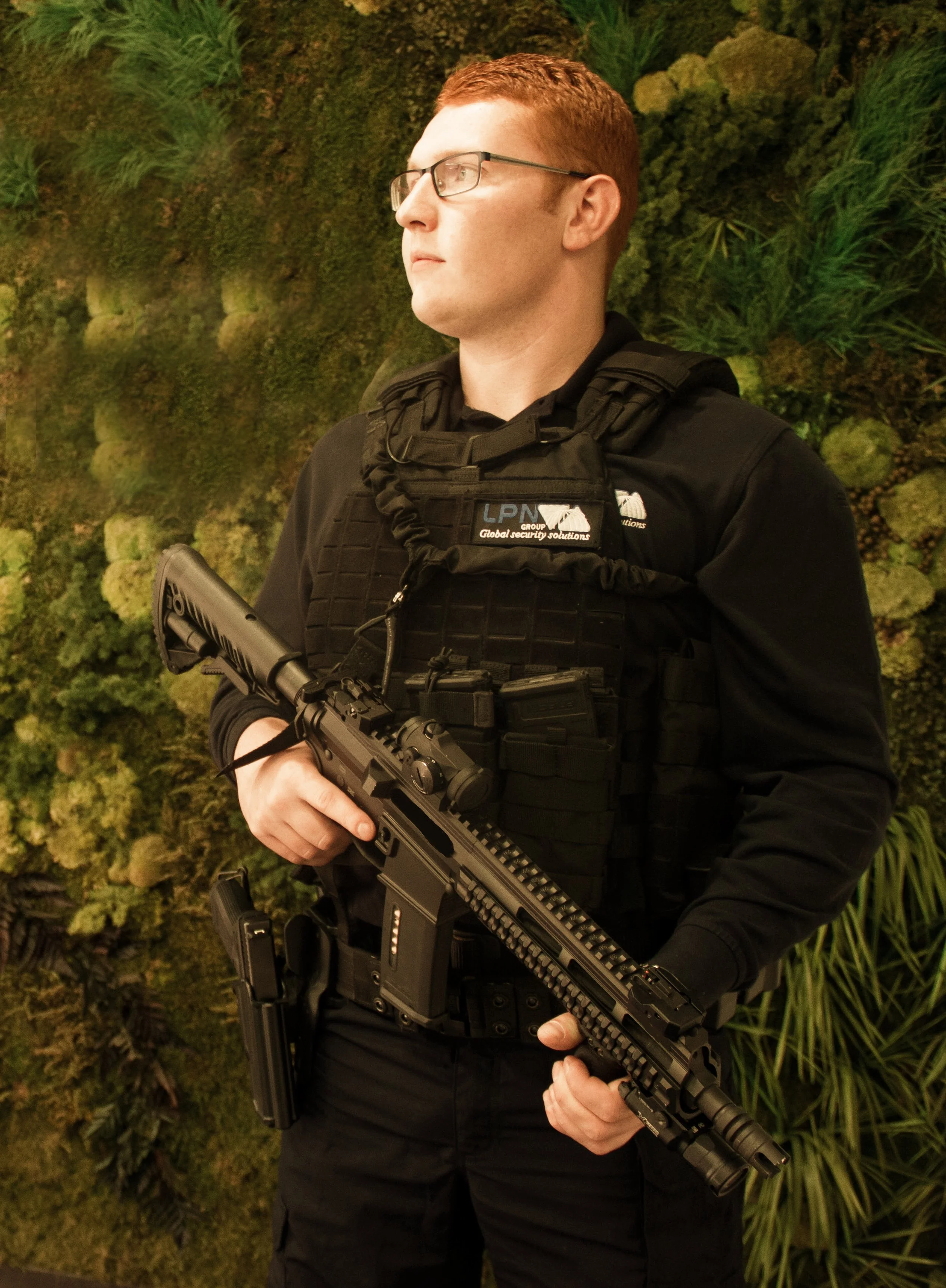 Un homme en tenue de sécurité armée, portant un fusil, portant un gilet pare-balles et des lunettes, se tient devant un mur végétal vert.