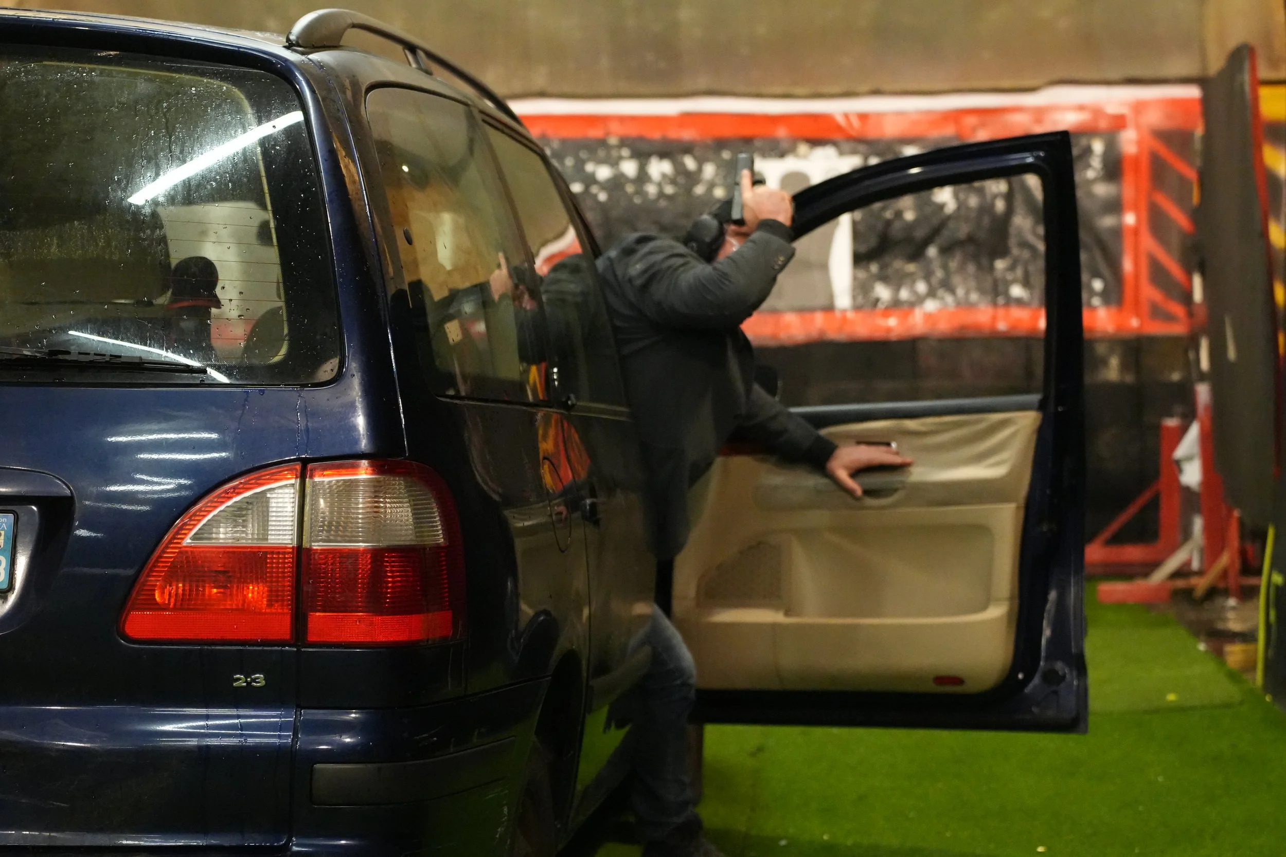 Un stagiaire en protection armée sortant d'une voiture noire, tenant une arme, à l'intérieur d'un bâtiment ou d'un garage, avec des équipements de sécurité visibles en arrière-plan.