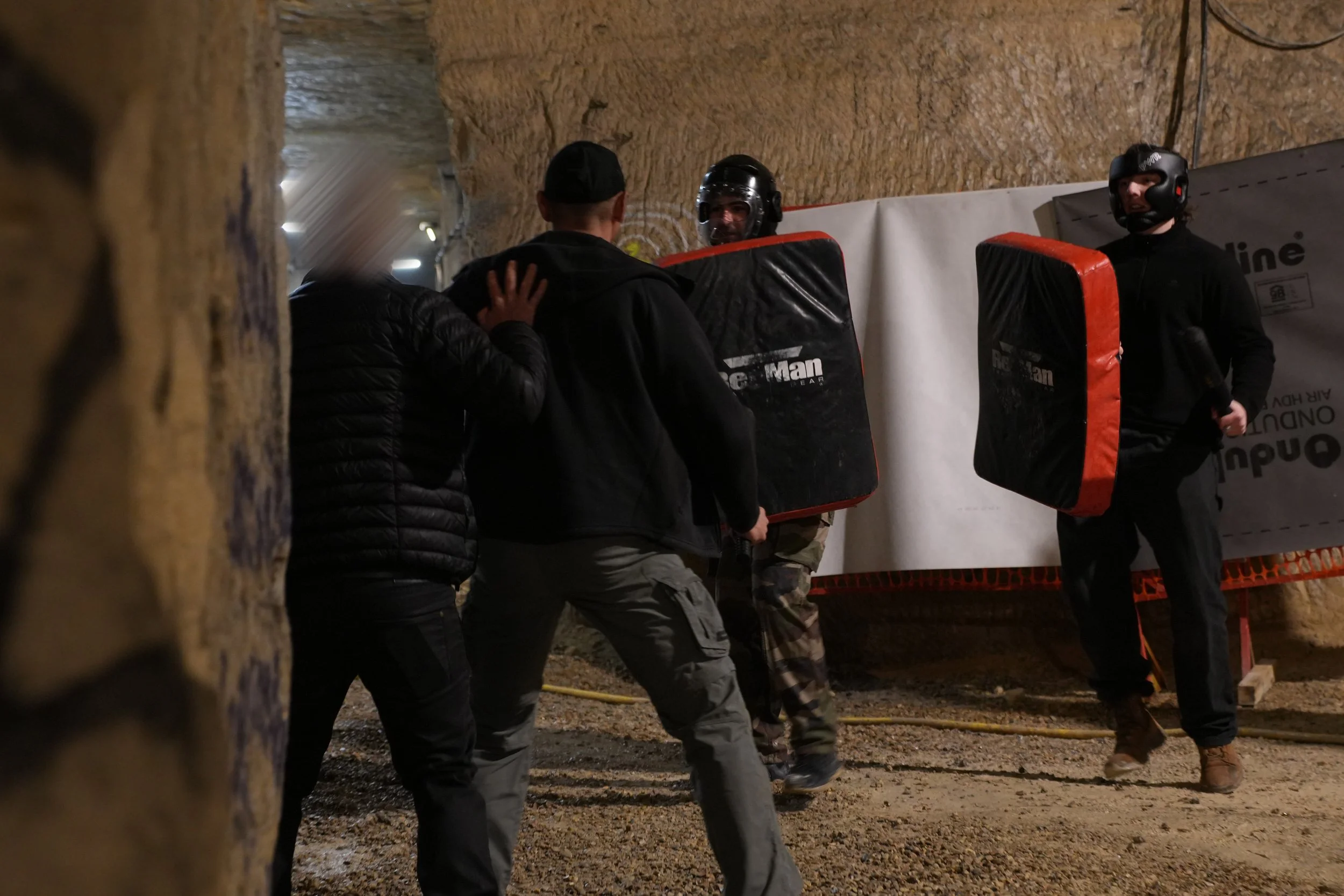 Deux personnes en tenue de protection maniant des boucliers pour se défendre contre une autre personne dans une cave ou tunnel.