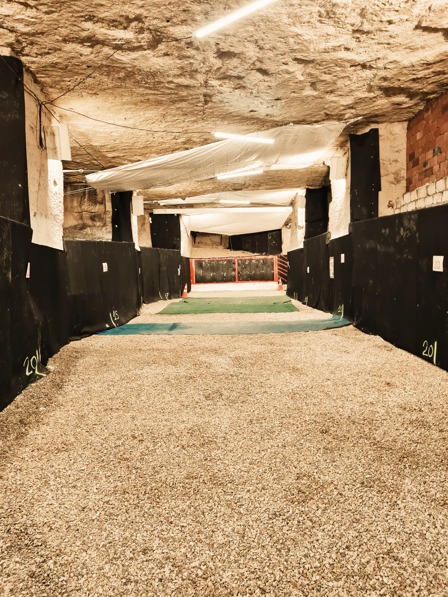 Tunnel ou pas de tir en sous sol avec un sol en gravier, entouré de barrières noires avec des numéros, éclairé par des lumières fluorescent, et en partie couvert de tissus blancs.