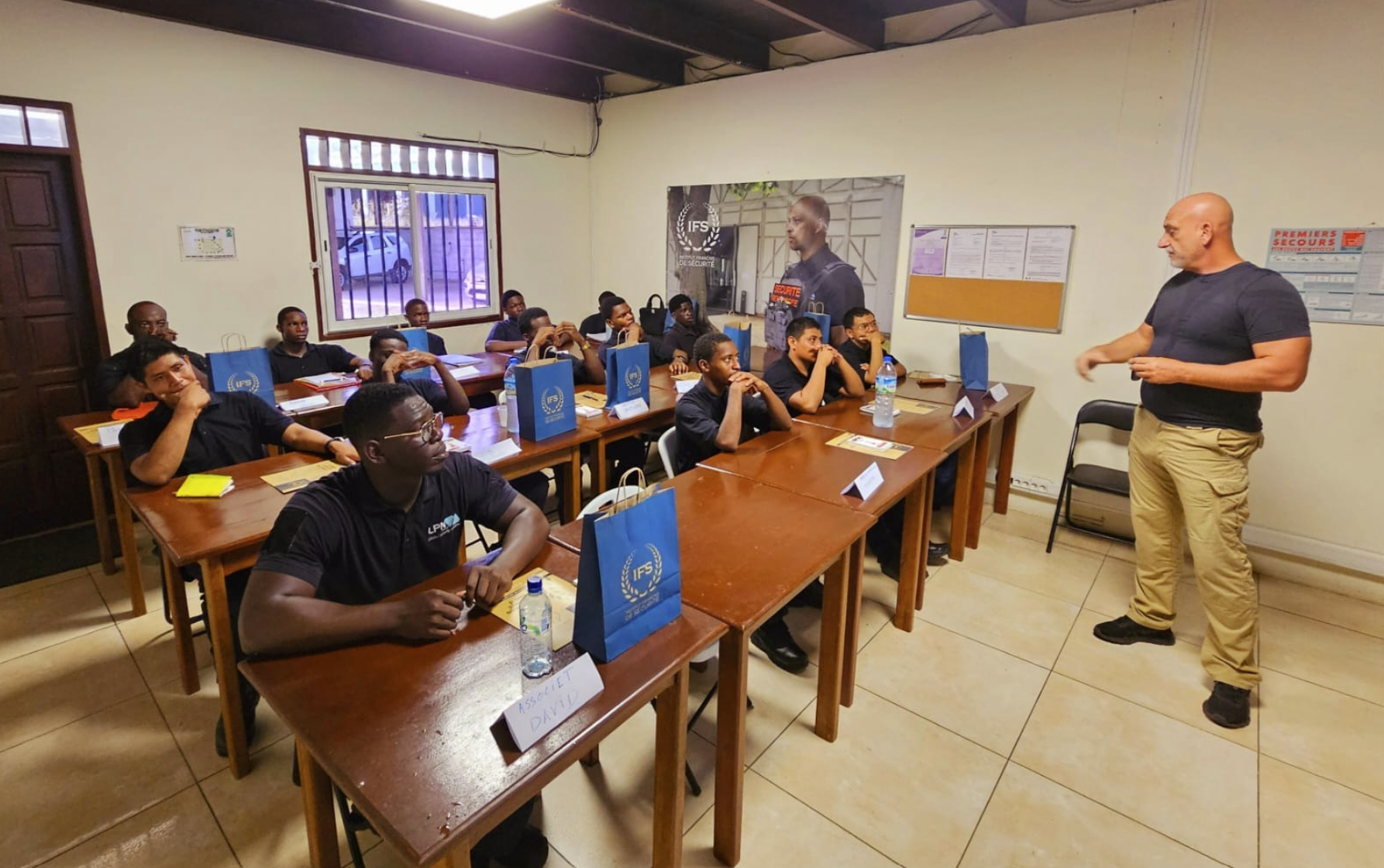 Un groupe d'étudiants en sécurité armée assis dans une salle de classe, en train d'écouter un instructeur devant un tableau blanc, avec des sacs bleus portant le logo IFS sur la table.