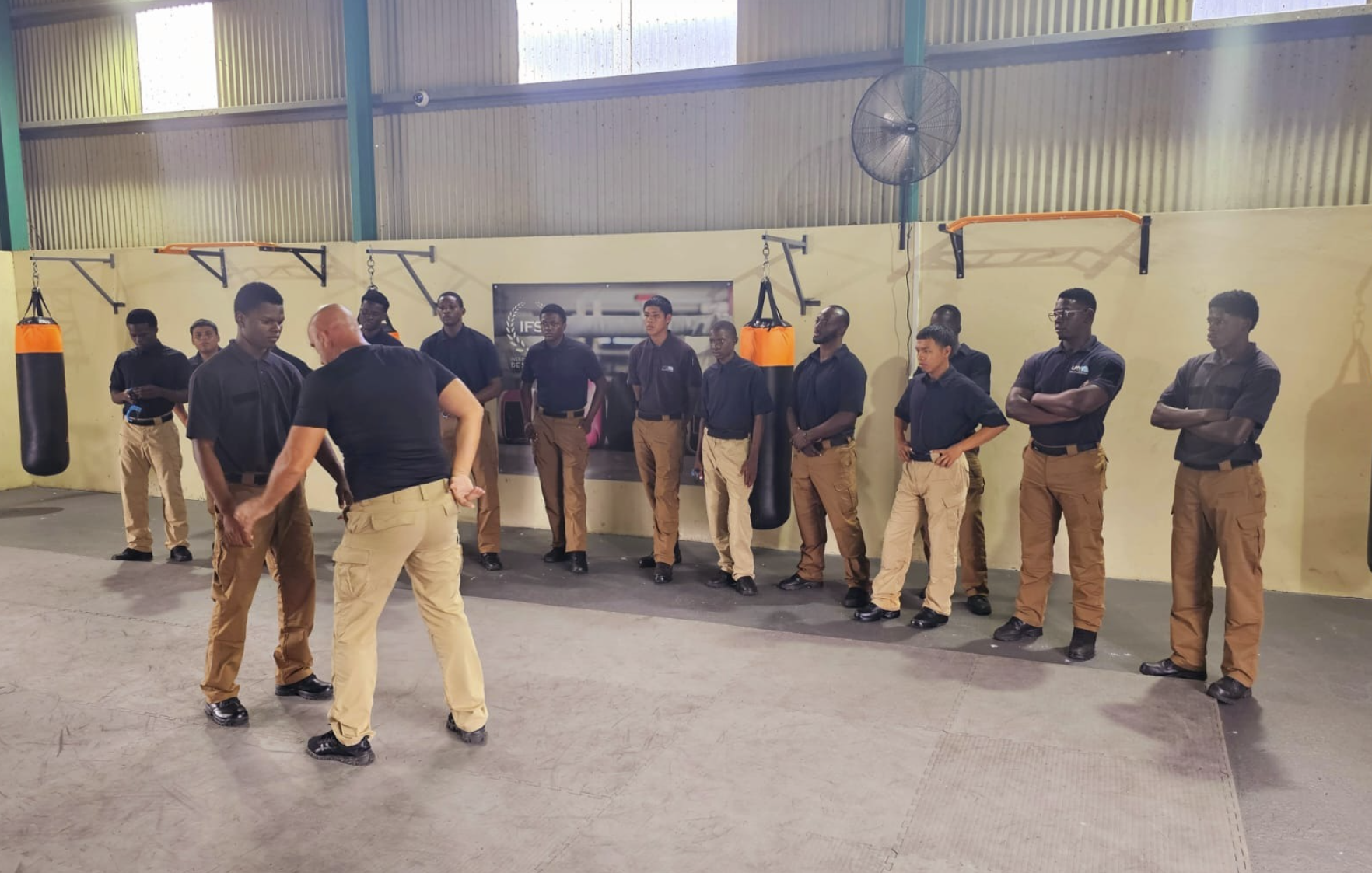 Instructeur en sécurité privée armée donnant des instructions à un groupe de jeunes stagiaires dans un dojo avec sacs de frappe