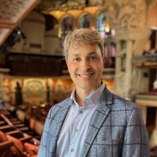 Ein lächelnder Mann in einem blauen Hemd und Blazer vor einer mit bunten Fenstern geschmückten Kirche