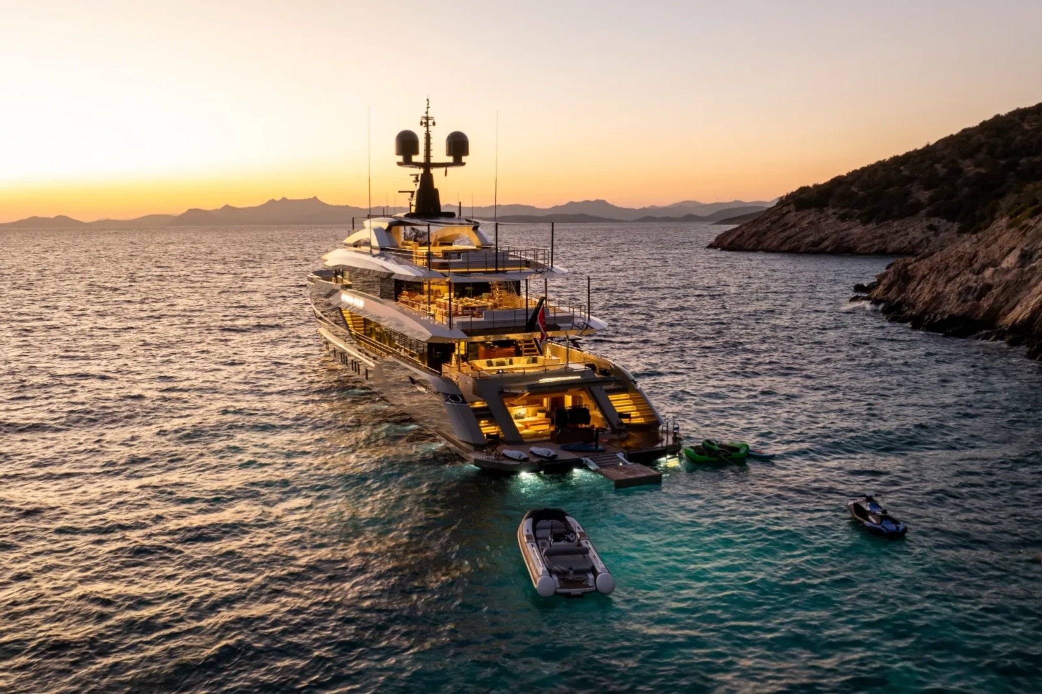 Luxury yacht illuminated at sunset with small boats floating nearby, surrounded by calm sea and distant hills.